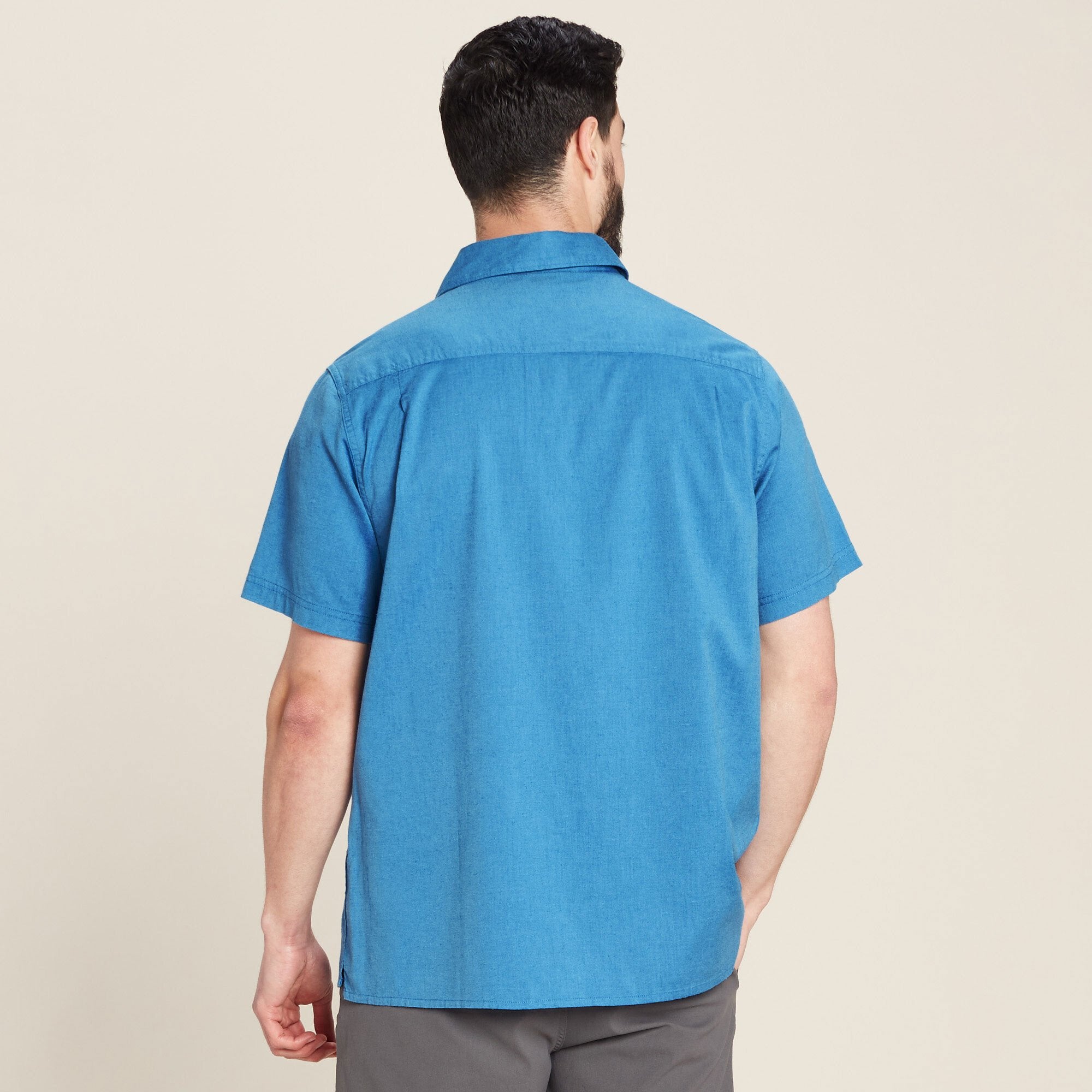 Rear view of the same Sherpa Adventure Gear Tharu Journey Short Sleeve Shirt in Blue. The back of the shirt is plain, without additional design elements, showcasing its clean-cut structure and simple tailoring. The model’s arms are down, and the shirt drapes naturally, illustrating the fit and length.