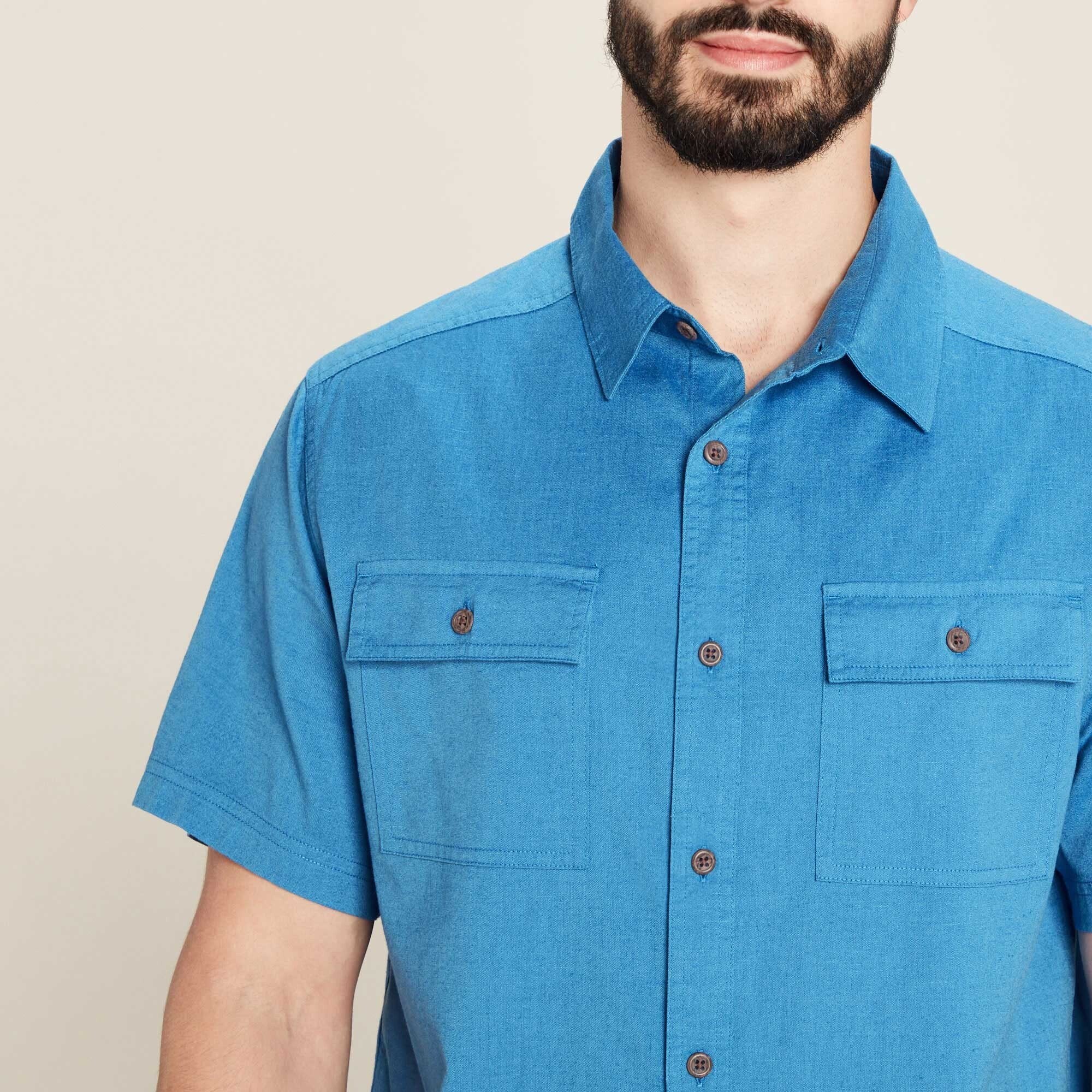 A close-up of the Sherpa Adventure Gear Tharu Journey Short Sleeve Shirt in Blue’s chest area, highlighting the intricate details of the buttoned chest pockets. The buttons are dark brown, adding a subtle contrast to the blue fabric. The texture of the shirt is slightly visible, showing a fine weave that gives the material a soft, lightweight appearance. The model’s beard and jawline frame the image, keeping the focus on the garment.