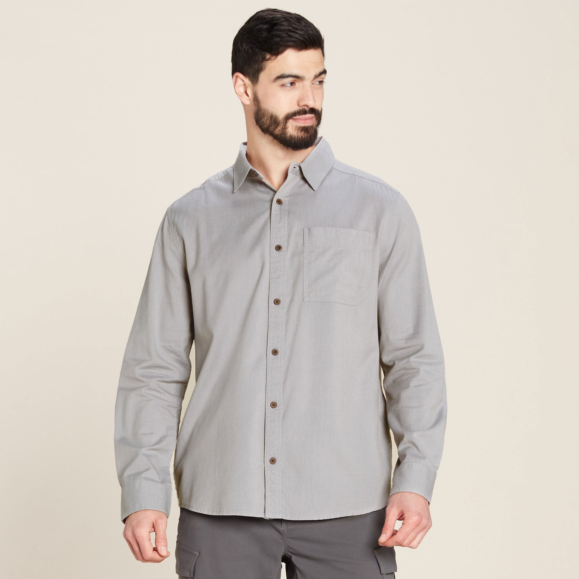 A man wearing a Sherpa Adventure Gear Tharu Long Sleeve Shirt in Grey with a single chest pocket, standing confidently and looking off to the side. The shirt features button details down the front and has a classic collar. The model pairs the shirt with dark grey pants.