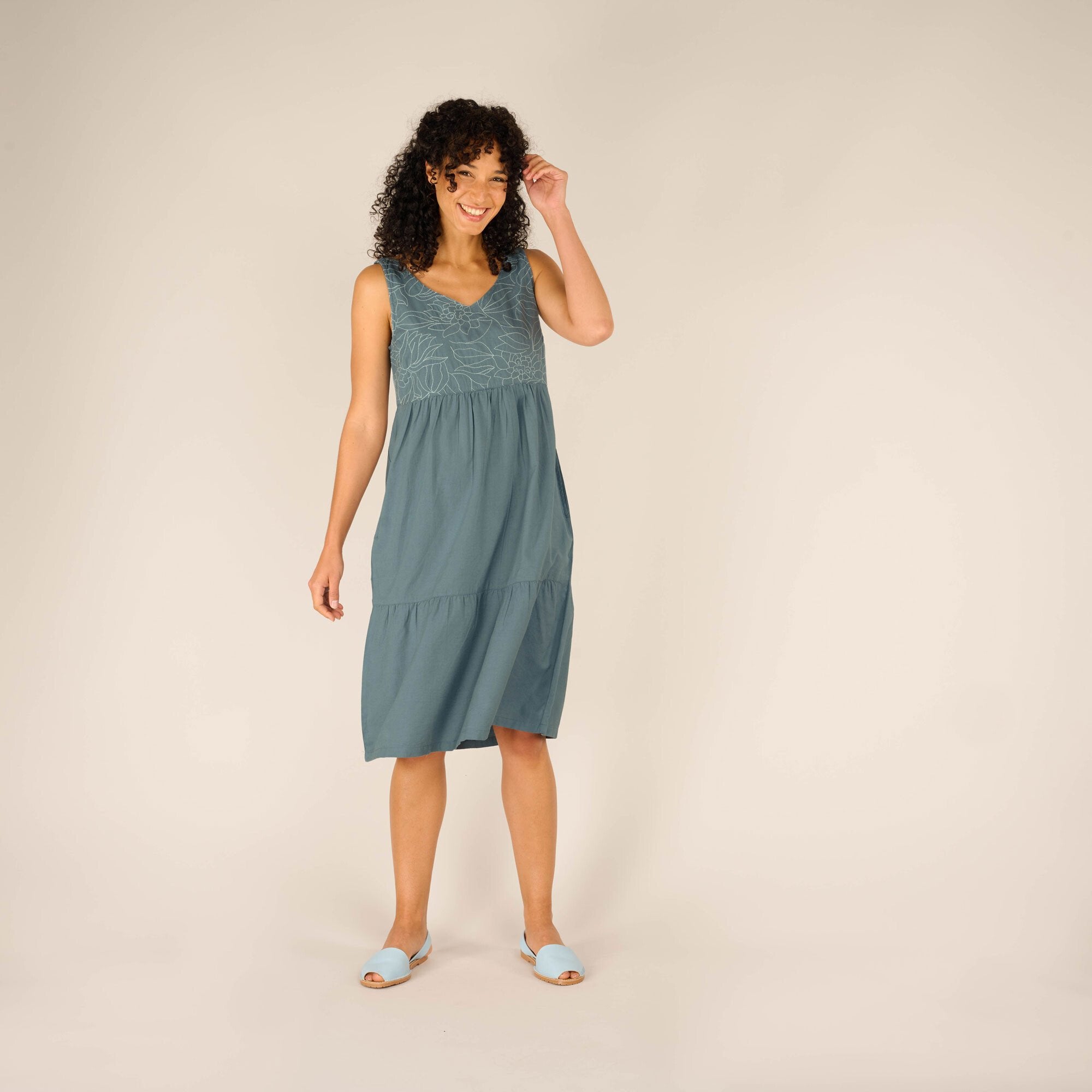 A woman with curly black hair and a smile is wearing a Sherpa Adventure Gear Tharu Tank Dress in Blue with floral embroidery on the upper chest area. The dress has a V-neckline, a relaxed fit, and falls just below the knees. She is standing with one hand adjusting her hair and the other relaxed at her side, wearing light blue open-toe sandals. The background is a neutral beige.