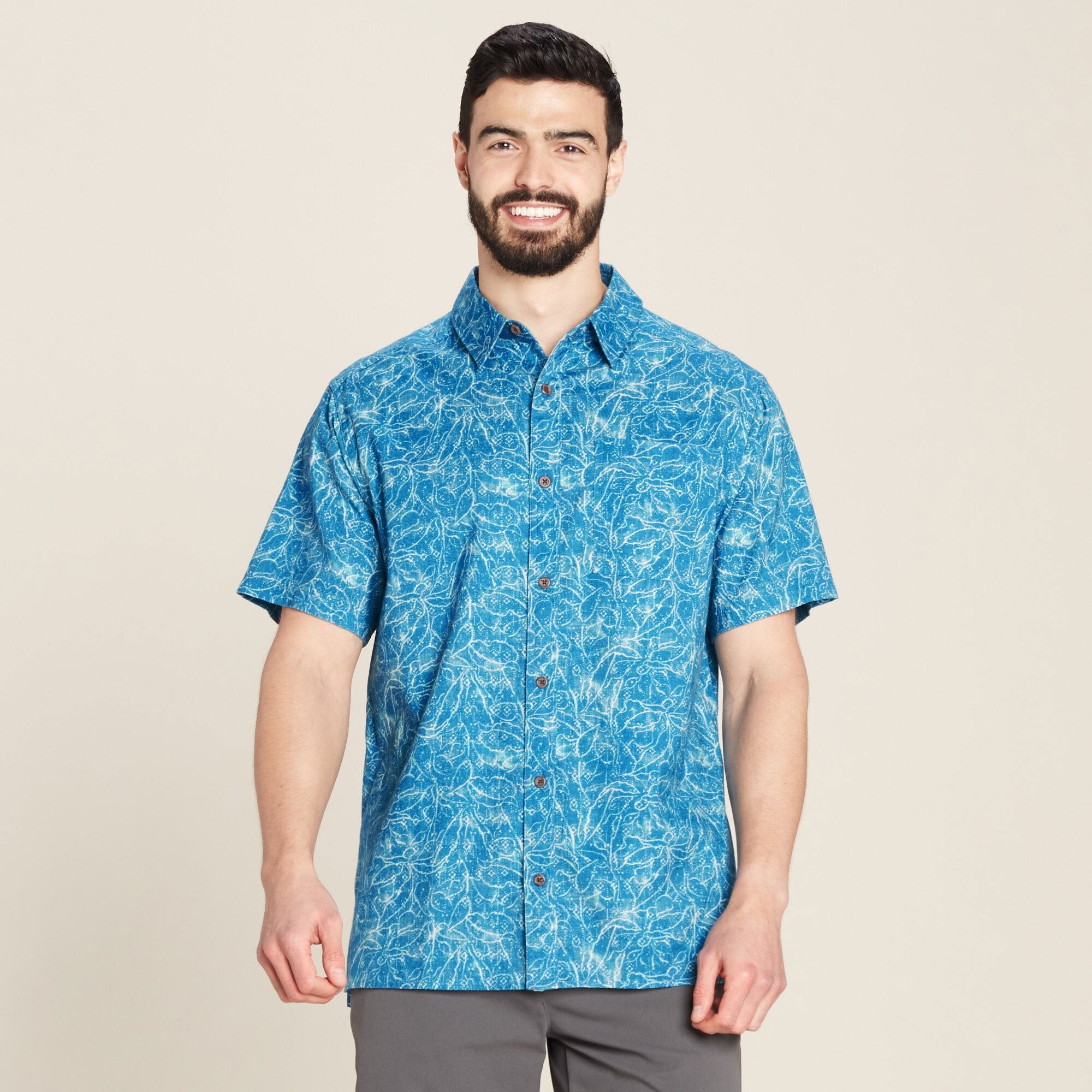 The man is smiling, showcasing the front of the Sherpa Adventure Gear Tharu Voyage Short Sleeve Shirt in Blue featuring a white botanical pattern. The shirt has a classic collar, short sleeves, and a button-down closure with visible wooden buttons. The lightweight fabric gives a relaxed and airy appearance. He pairs the shirt with grey pants.