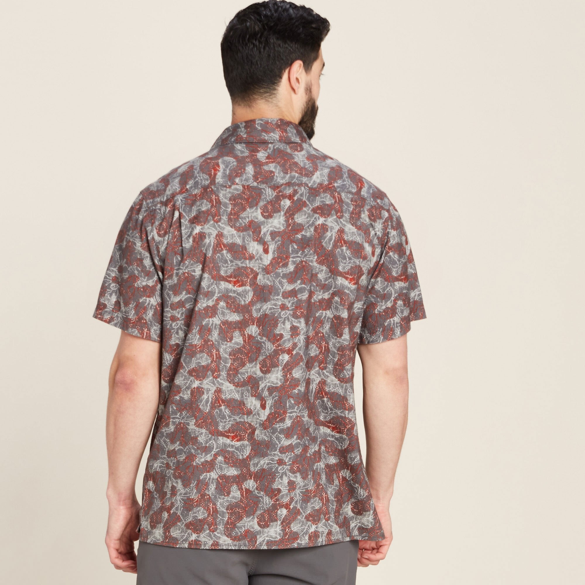 Rear view of the same Sherpa Adventure Gear Tharu Voyage Short Sleeve Shirt in Grey, highlighting the seamless back design and continuation of the abstract red and grey print. The model stands straight, with the shirt untucked, emphasising its casual fit and straight hemline.