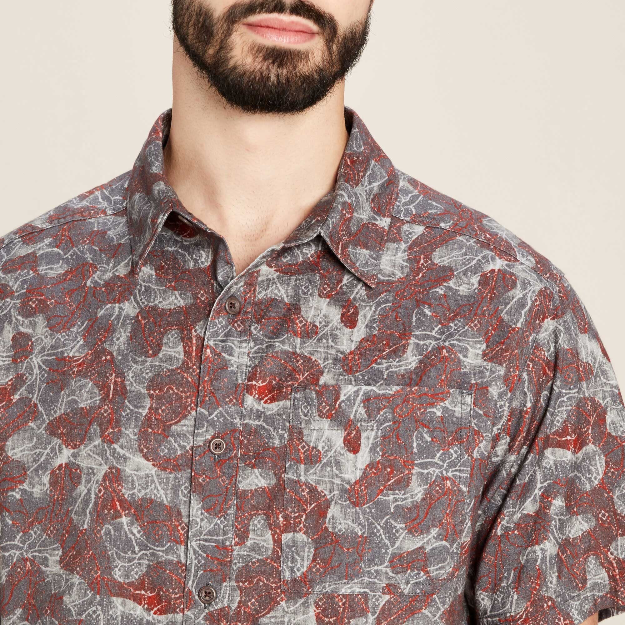 A close-up view of the Sherpa Adventure Gear Tharu Voyage Short Sleeve Shirt in Grey's upper front portion, capturing the intricate pattern of red and grey tones in greater detail. The buttons and collar structure are clearly visible, as is the single chest pocket blending into the abstract print. This image showcases the craftsmanship and fabric texture.