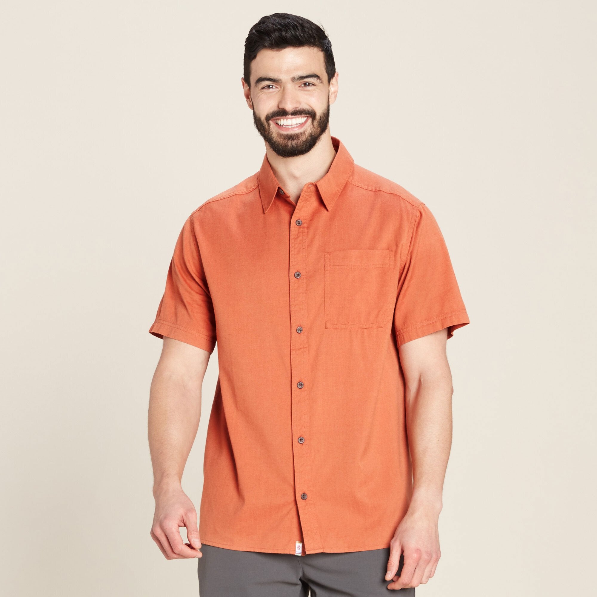 A man is wearing a Sherpa Adventure Gear Tharu Voyage Short Sleeve Shirt in Orange, button-down shirt in a warm orange shade. The shirt features a front pocket on the left side, adding functionality to the minimalistic design. The fabric appears lightweight and breathable, ideal for warm weather. The model is smiling, giving a friendly and casual vibe.