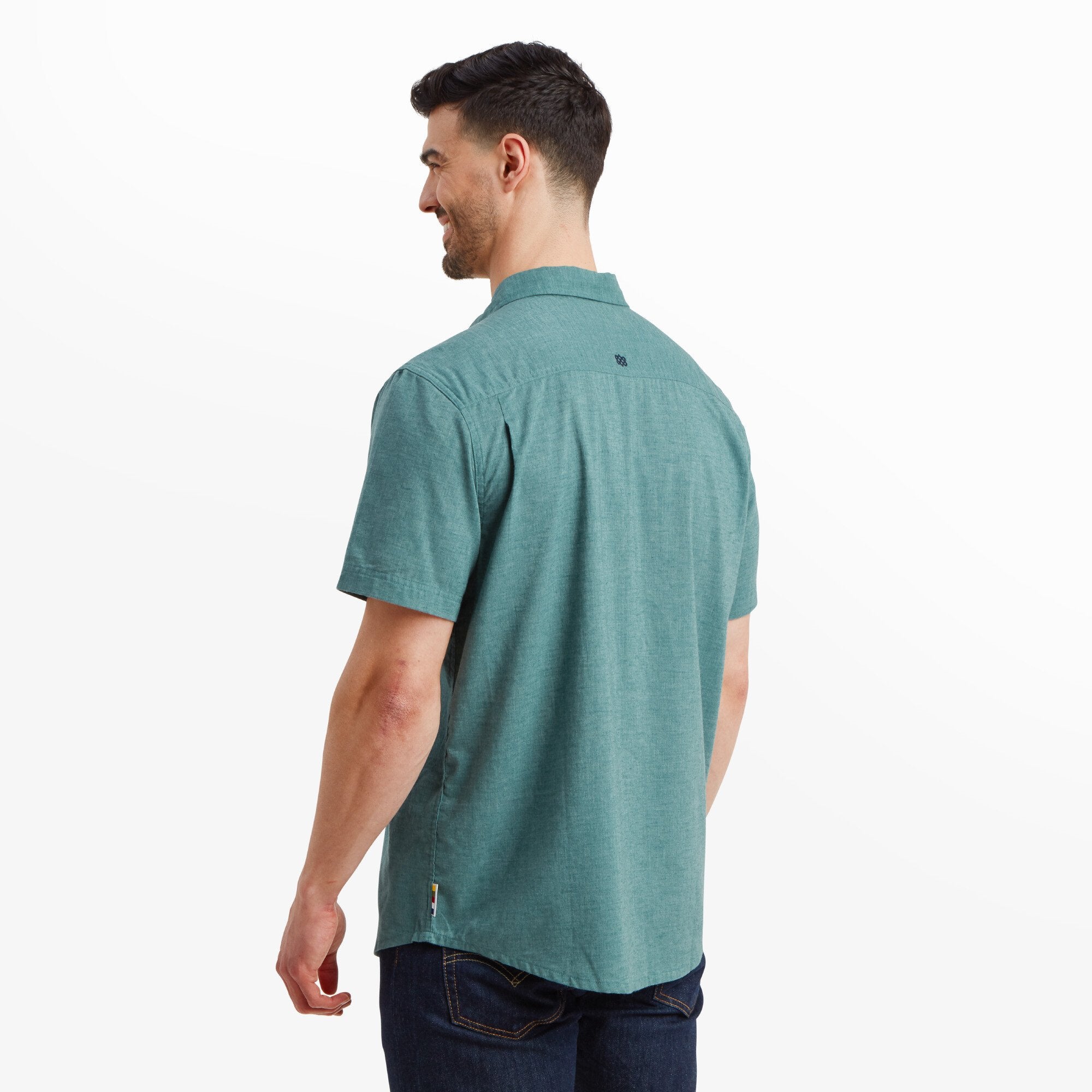 The back view of the Sherpa Adventure Gear Tibet Stripe Short Sleeve Shirt in Blue, emphasising the solid teal base with a subtle logo embroidery at the upper back. The shirt flows smoothly over the shoulders, with the model's arms relaxed by his sides.