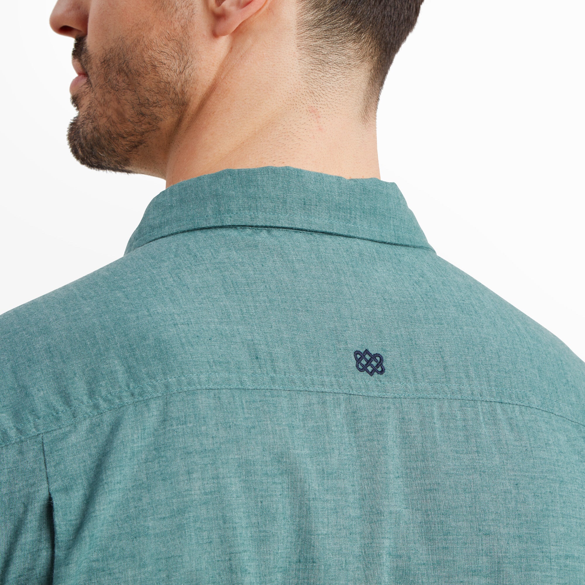 A zoomed-in view of the back neckline of the Sherpa Adventure Gear Tibet Stripe Short Sleeve Shirt in Blue, showcasing the small embroidered logo in dark navy. The close-up highlights the fine texture of the fabric and the gentle pleating at the back yoke for added comfort and style.