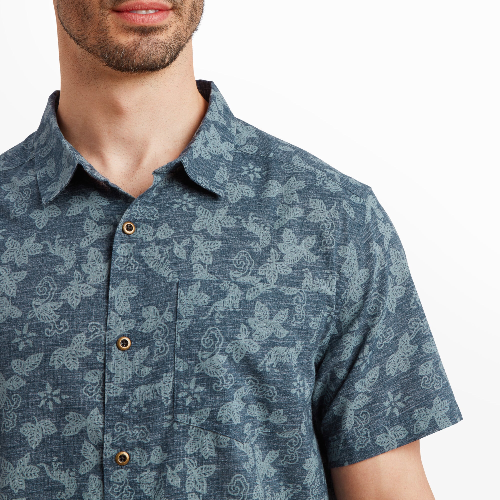 A close-up of the Sherpa Adventure Gear Tiger Floral Short Sleeve Shirt in Blue's front chest area, focusing on the floral pattern and button-up design. The fabric texture is highlighted, and the single front pocket is visible, demonstrating the fine details and craftsmanship of the garment. The model's collar is slightly open, adding to the casual aesthetic.