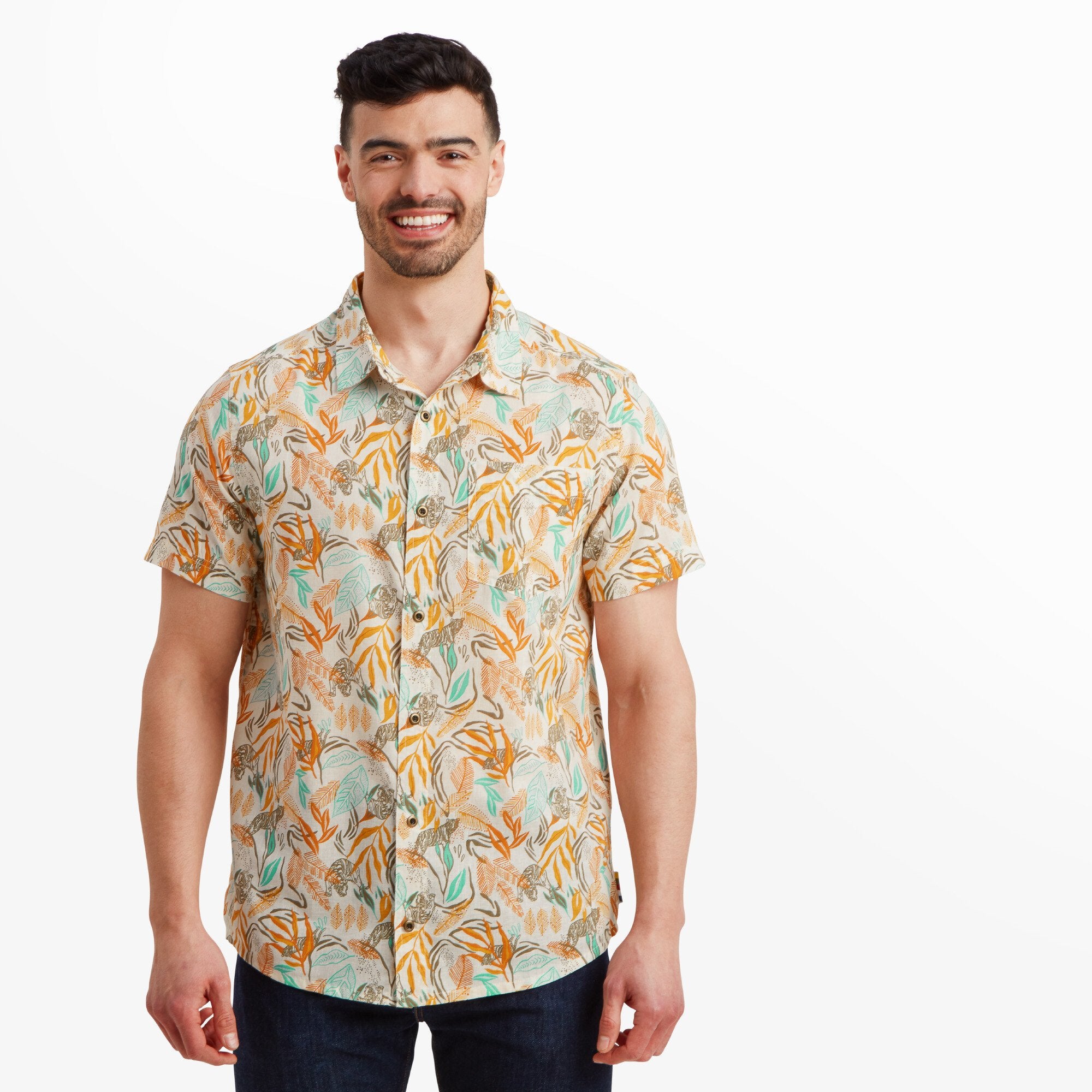 A smiling man wearing a Sherpa Adventure Gear Tiger Leaf Short Sleeve Shirt in Orange featuring a vibrant orange and beige leaf and tiger print. The shirt is worn untucked, paired with dark denim jeans.