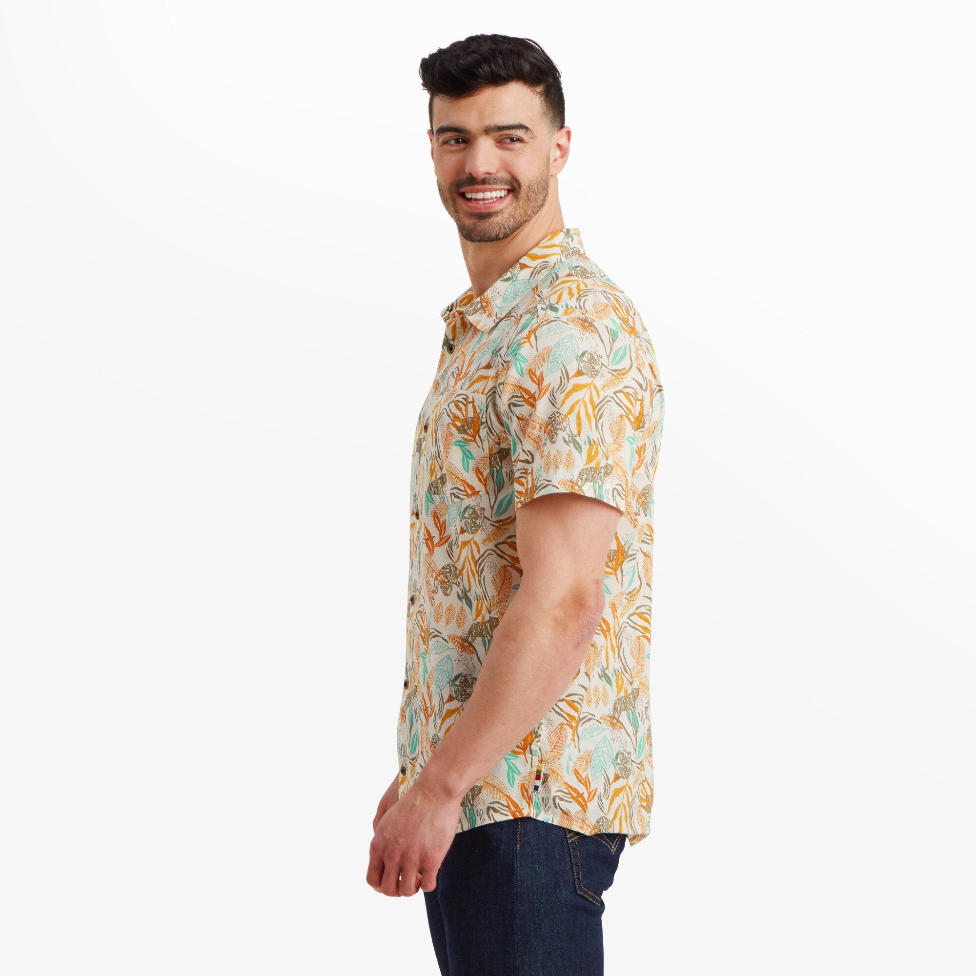 The man is captured from a side angle, showcasing the Sherpa Adventure Gear Tiger Leaf Short Sleeve Shirt in Orange's relaxed fit and hemline. The tropical pattern remains consistent along the shirt, and the man has a bright smile.