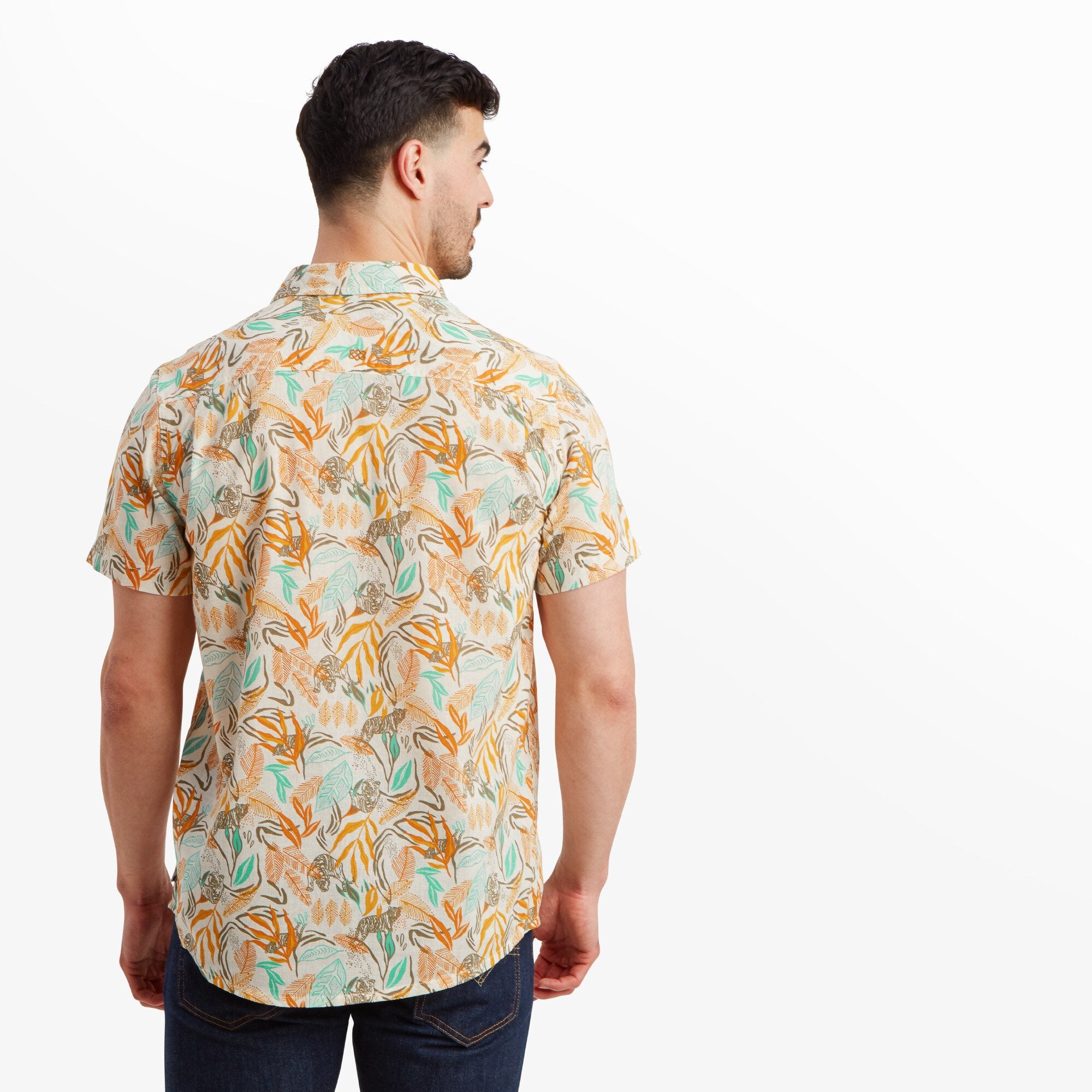 A rear view of the Sherpa Adventure Gear Tiger Leaf Short Sleeve Shirt in Orange highlighting the pattern continuity across the back, with clean stitching and a slightly curved hemline. The man pairs the shirt with dark jeans for a casual look.