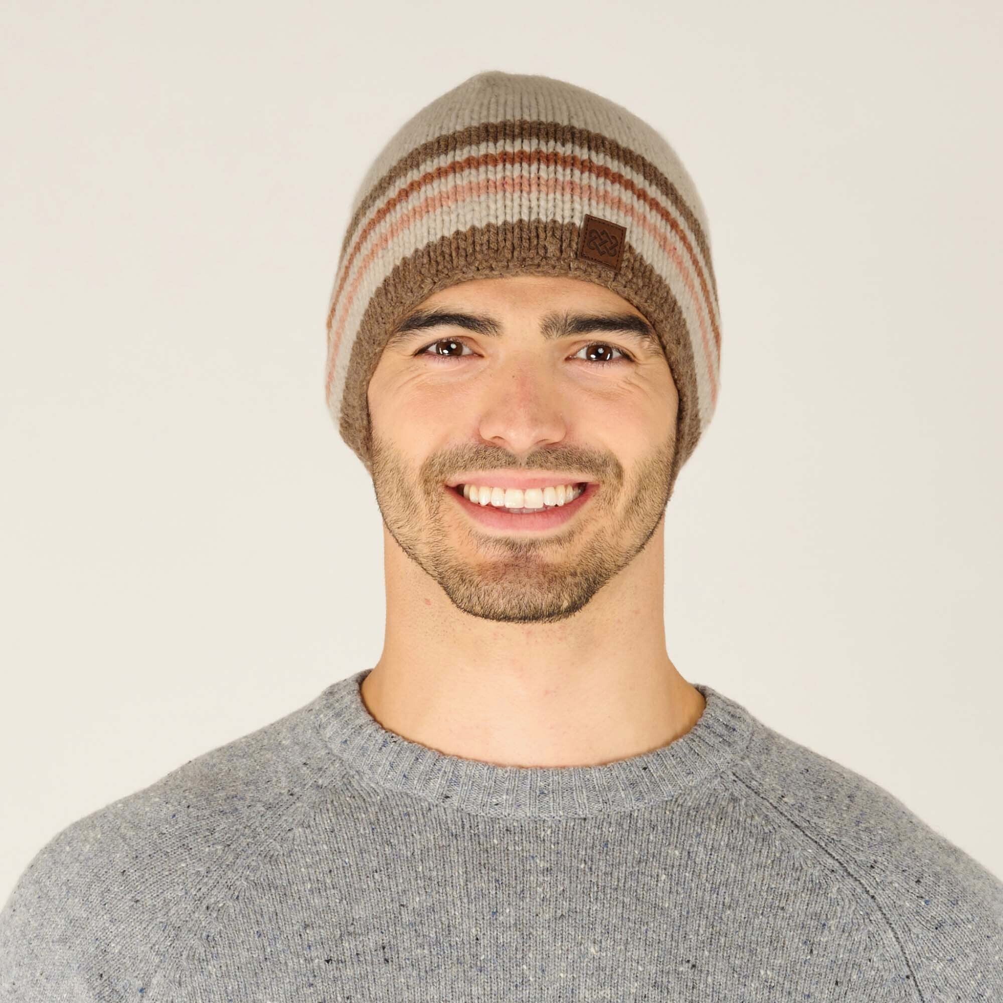 A close-up shot of a smiling model wearing a Sherpa Adventure Gear Tsho Stripe Hat in Beige with horizontal stripes in brown and pinkish hues. The hat features a small rectangular leather patch with the Sherpa logo, positioned at the front. The model is wearing a light gray speckled sweater, complementing the neutral tones of the hat.