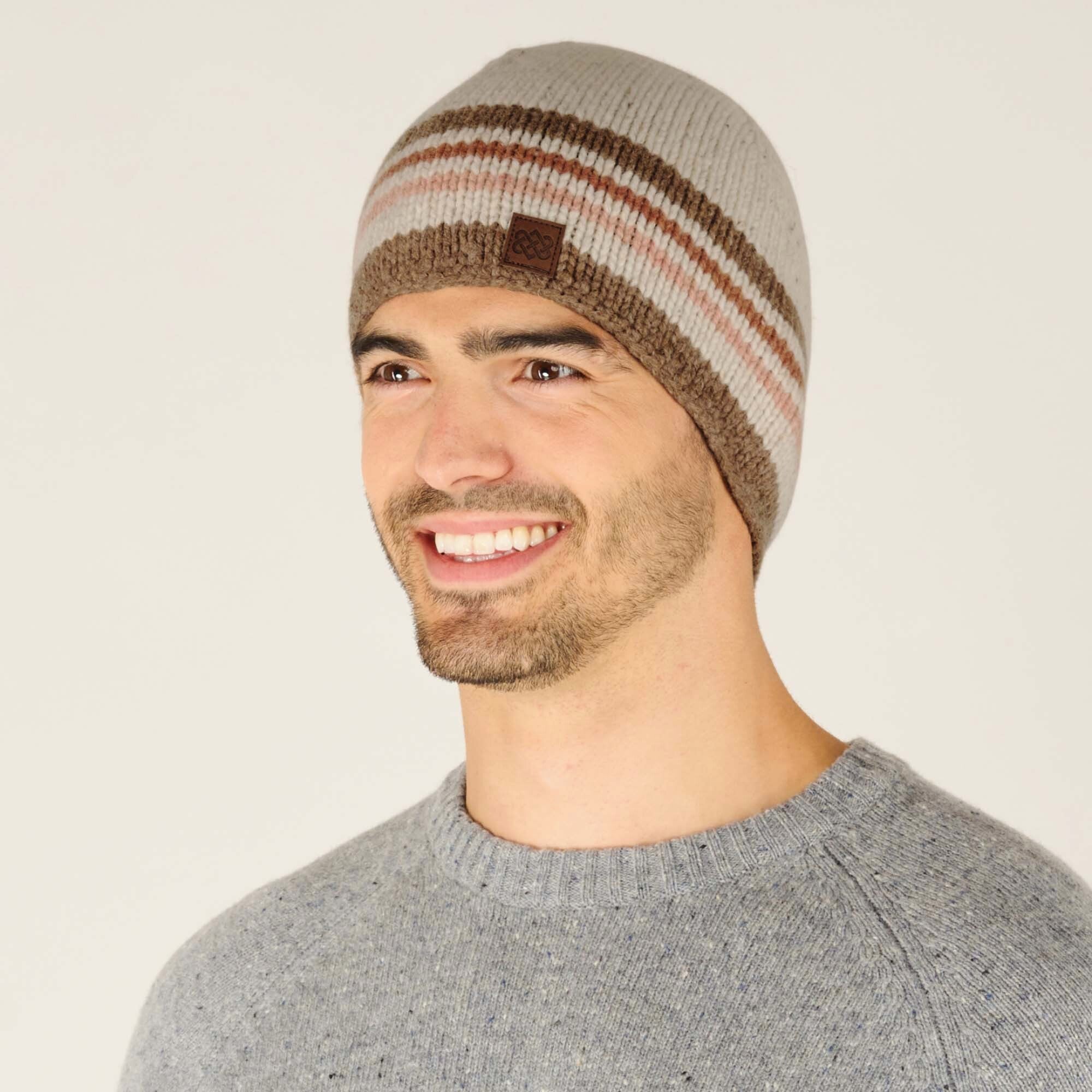 A profile view of the same Sherpa Adventure Gear Tsho Stripe Hat in Beige, showcasing its snug fit and the alignment of the stripes across the head. The Sherpa logo patch is visible on the side, and the model's friendly expression highlights the comfortable and stylish design. The model's light gray sweater subtly accentuates the earthy color palette of the hat.