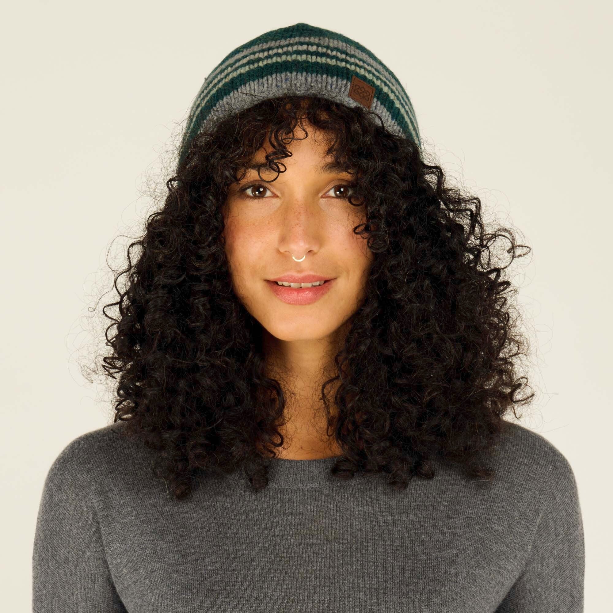 A woman with curly black hair wearing a Sherpa Adventure Gear Tsho Stripe Hat in Green in shades of green and gray. The beanie features a small leather patch with a logo on the side. She is smiling softly, wearing a gray sweater, and is photographed against a neutral background.