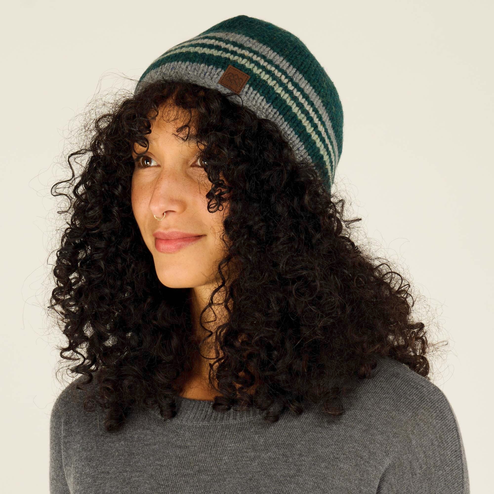 Side profile of the same woman with curly black hair wearing the Sherpa Adventure Gear Tsho Stripe Hat in Green. The leather patch logo is visible, and she is smiling slightly while wearing a gray sweater. The background remains neutral to highlight the hat.