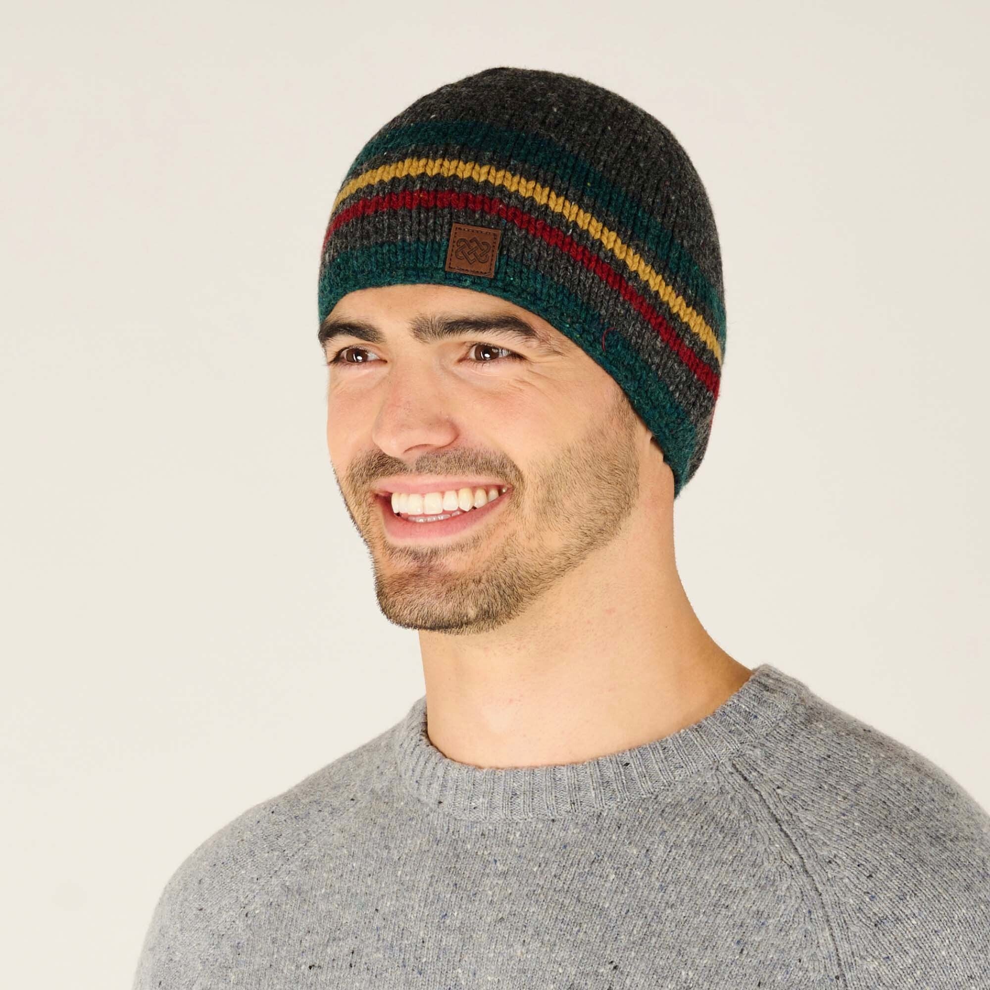 The same man is shown from a three-quarter angle, giving a better view of the side of the Sherpa Adventure Gear Tsho Stripe Hat in Grey. The beanie's colors—dark green, mustard yellow, burgundy red, and charcoal grey—wrap around the head, and the leather logo patch is still visible on the front. The man is smiling and looking slightly to the side. His heather grey sweater with speckled details is also visible, and the plain off-white background remains consistent.