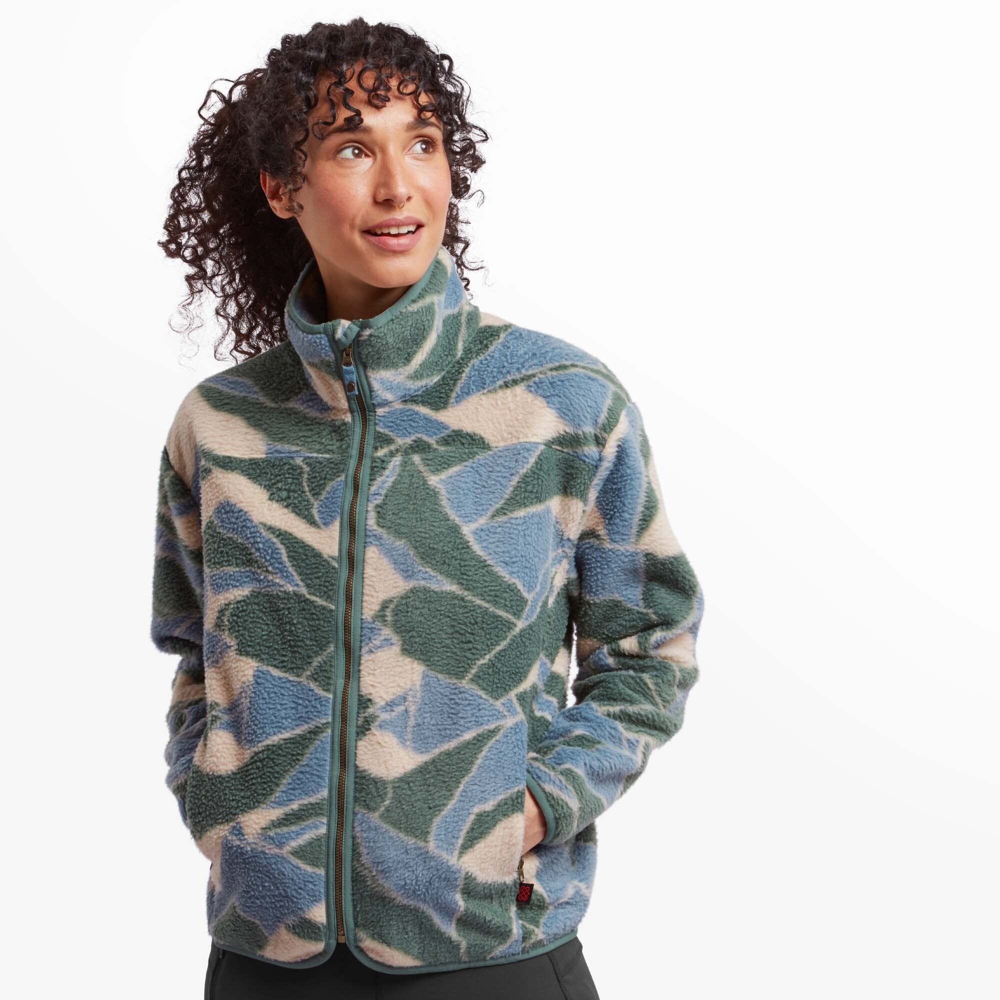 A woman with curly dark hair is wearing a Sherpa Adventure Gear Uddesya Eco Jacket in Blue with an abstract geometric pattern in shades of blue, green, and beige. The jacket has a high collar and a front zipper with a green trim. She is standing with her hands tucked in the pockets, looking off to the side with a pleasant expression. The background is white.