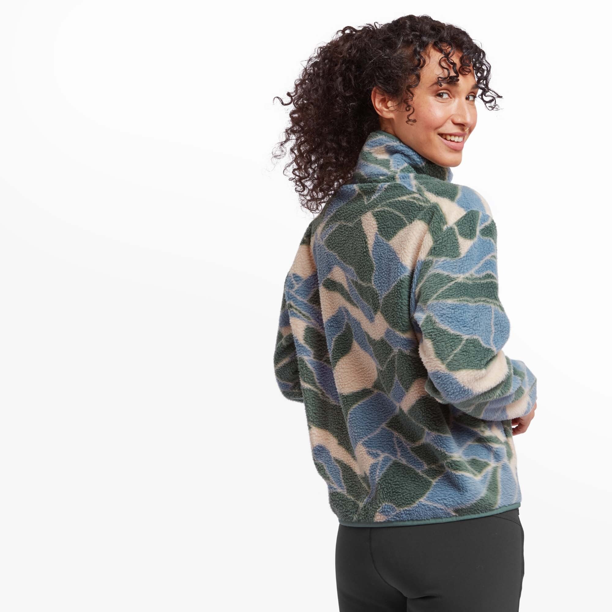 A rear view of the woman wearing the Sherpa Adventure Gear Uddesya Eco Jacket in Blue. She is turning her head slightly to smile at the camera. The abstract pattern continues across the back, and the collar remains upright. The jacket appears loose-fitting and warm.