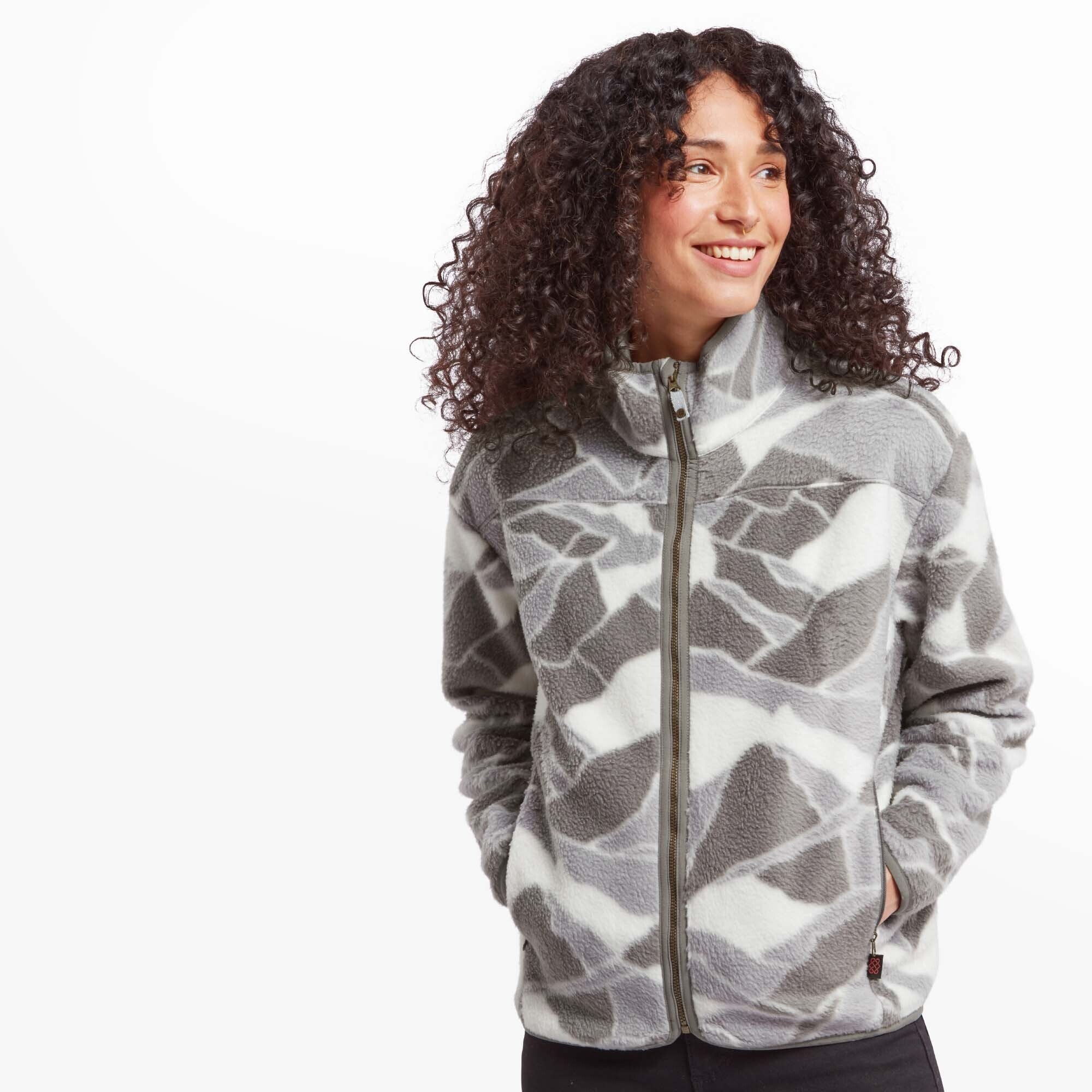 A woman with curly dark hair is wearing a Sherpa Adventure Gear Uddesya Eco Jacket in Grey with a geometric pattern. She stands slightly turned to her left, smiling while looking off-camera. The fleece jacket features a full-length zipper, a high collar, and two zippered pockets on either side. She has her hands tucked into the pockets, and the jacket’s cozy texture is visible.