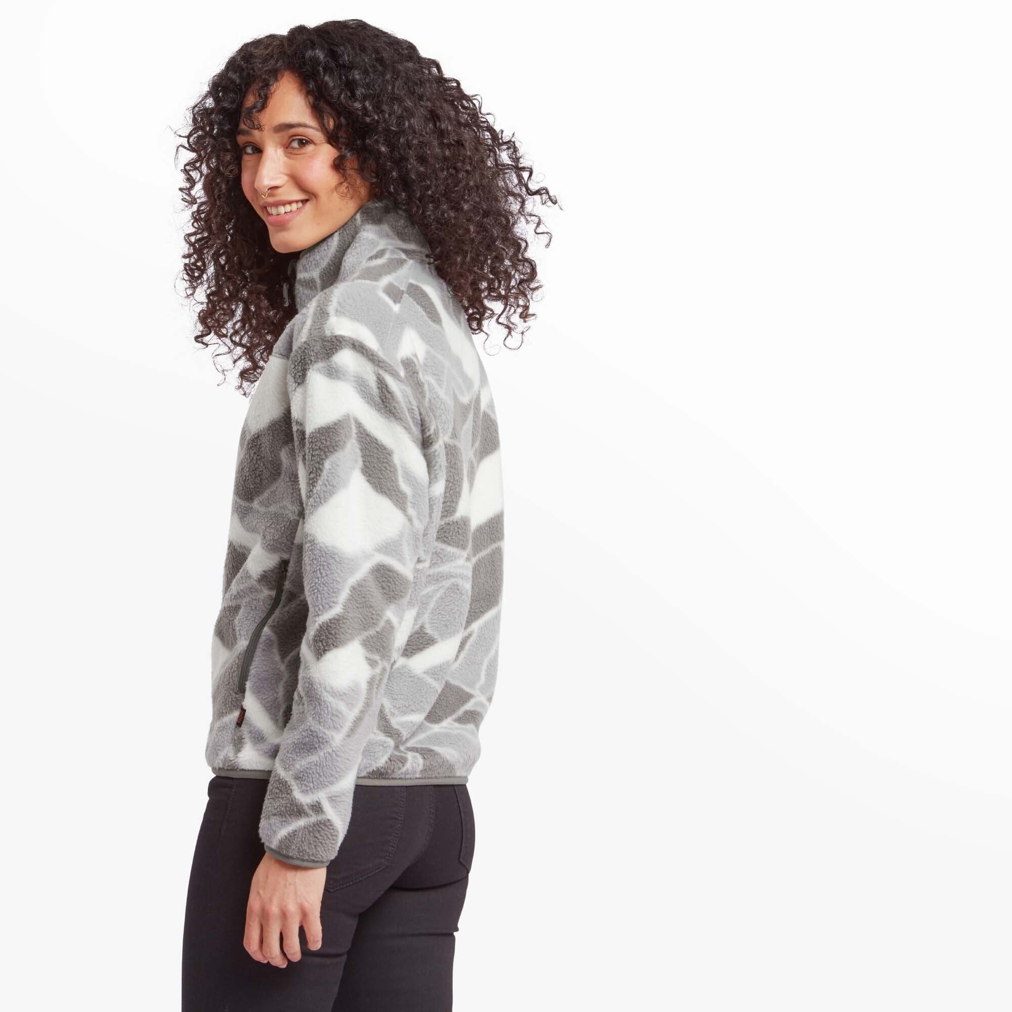 The model is captured from a three-quarter rear angle, smiling while turning her head towards the camera. The Sherpa Adventure Gear Uddesya Eco Jacket in Grey’s geometric pattern is showcased, along with the slightly elastic cuffs and hem. The fleece material appears soft and plush.