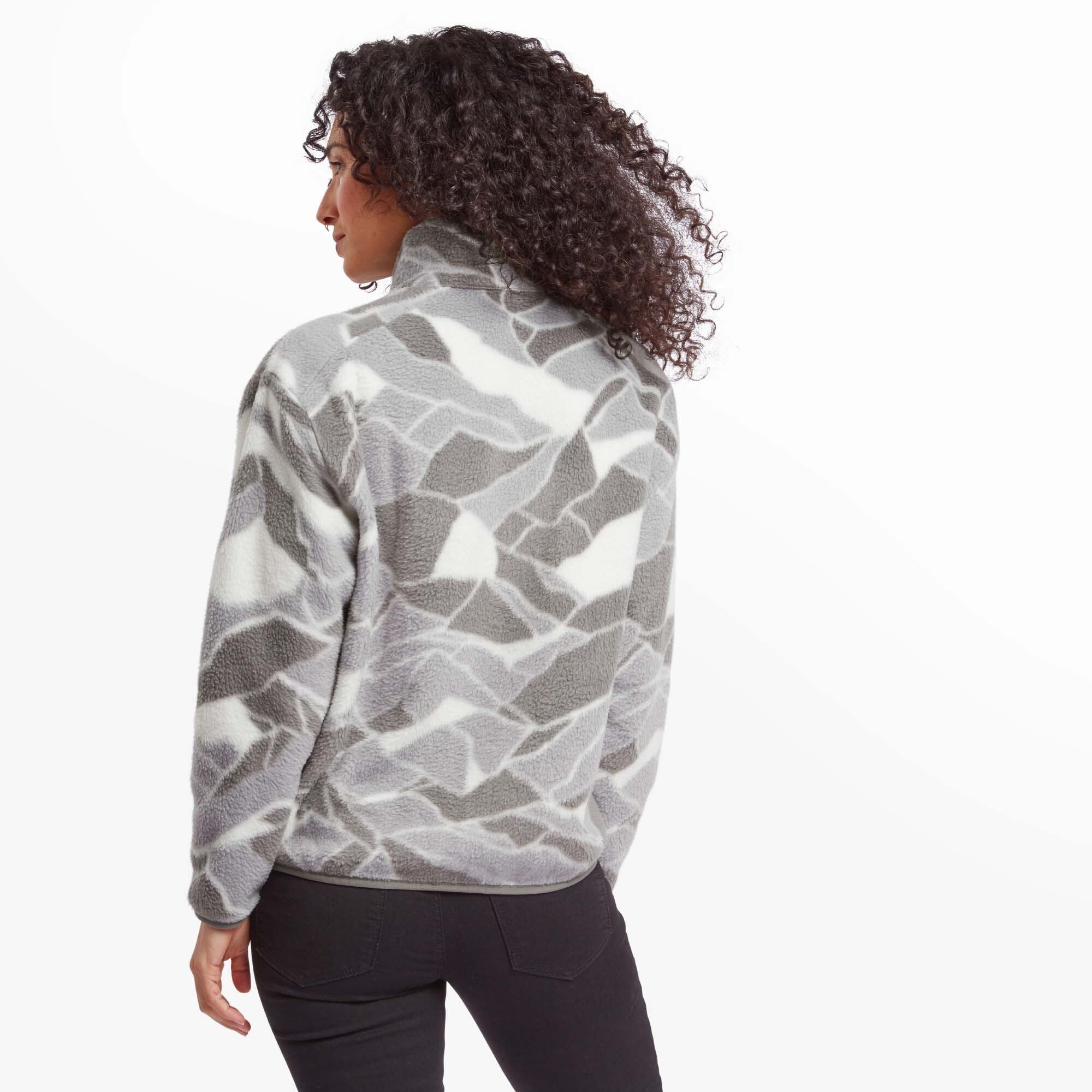 The back of the Sherpa Adventure Gear Uddesya Eco Jacket in Grey is visible as the model turns away from the camera. The jacket maintains the geometric pattern throughout, with a high collar for added warmth. Her curly hair cascades down her back, partially covering the collar.