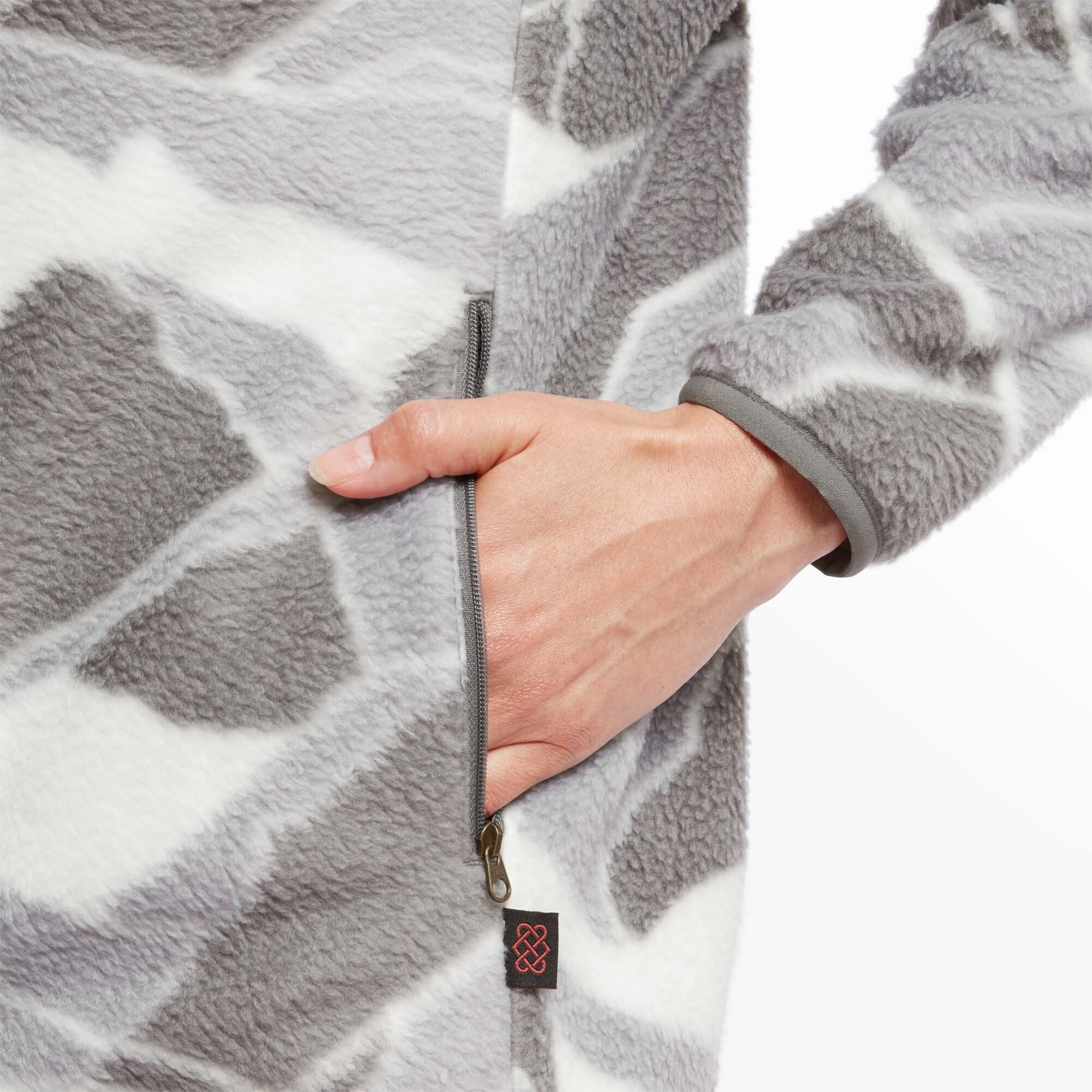 A close-up shot of the Sherpa Adventure Gear Uddesya Eco Jacket in Grey’s pocket area, showing the fleece texture and the zipper with a small pull tab. The model’s hand is partially inserted into the pocket, revealing the edge of the jacket’s cuff with its neatly finished trim. A small Sherpa Adventure Gear logo tag is stitched near the bottom hem.