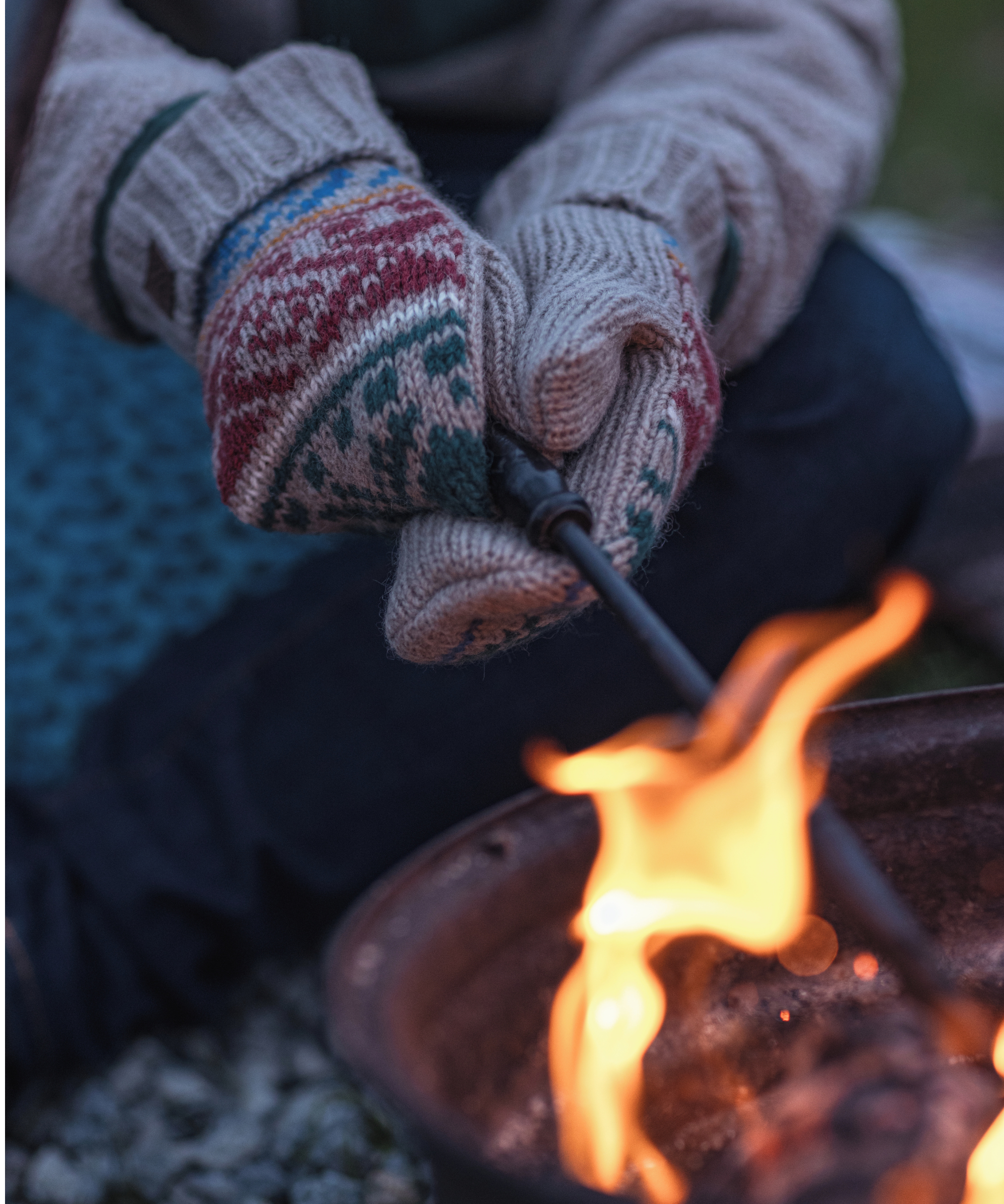 A close-up image of hands wearing cozy Sherpa Adventure Gear Hanssi Mittens in Beige with intricate red, green, and blue patterns near the wrists. The mittens are holding a metal fire starter, with visible sparks flying near a glowing campfire. The flames from the fire create a warm, flickering light in the foreground, contrasting against the dark background and highlighting the texture of the mittens. The scene evokes a sense of warmth and outdoor adventure.