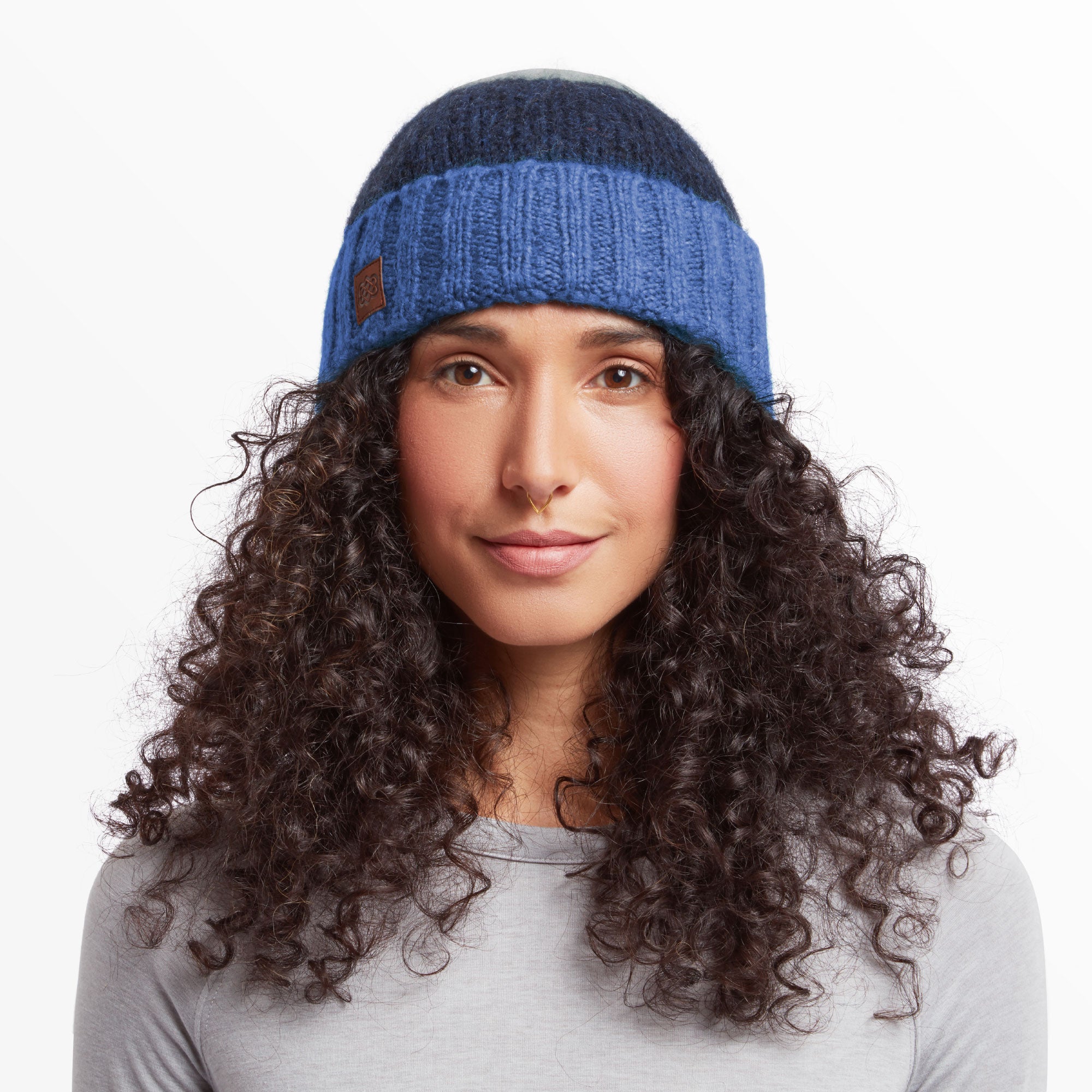 A close-up front view of a person wearing a Sherpa Adventure Gear Tenbu Hat in Blue. The hat has a ribbed design and features a small, square leather patch on the front. The individual has curly hair flowing out beneath the hat and is wearing a light gray top. Their expression is neutral yet soft, with minimal makeup highlighting their natural features.