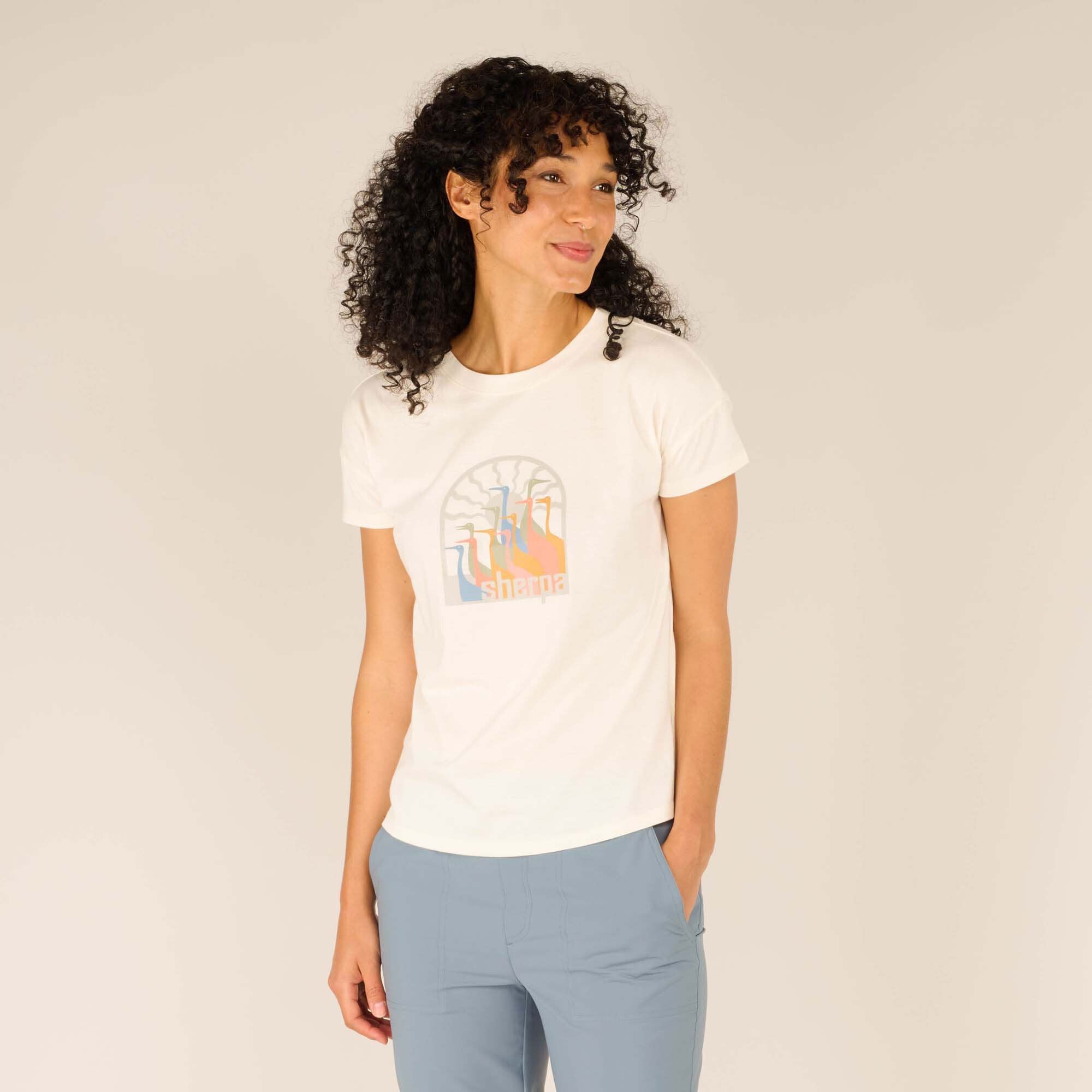 Front view of the Sherpa Adventure Gear Varuna Short Sleeve Crew in Cream worn by a smiling model. The t-shirt features a colorful Sherpa logo graphic on the chest, paired with light blue tailored pants, and the model’s curly hair frames her face.