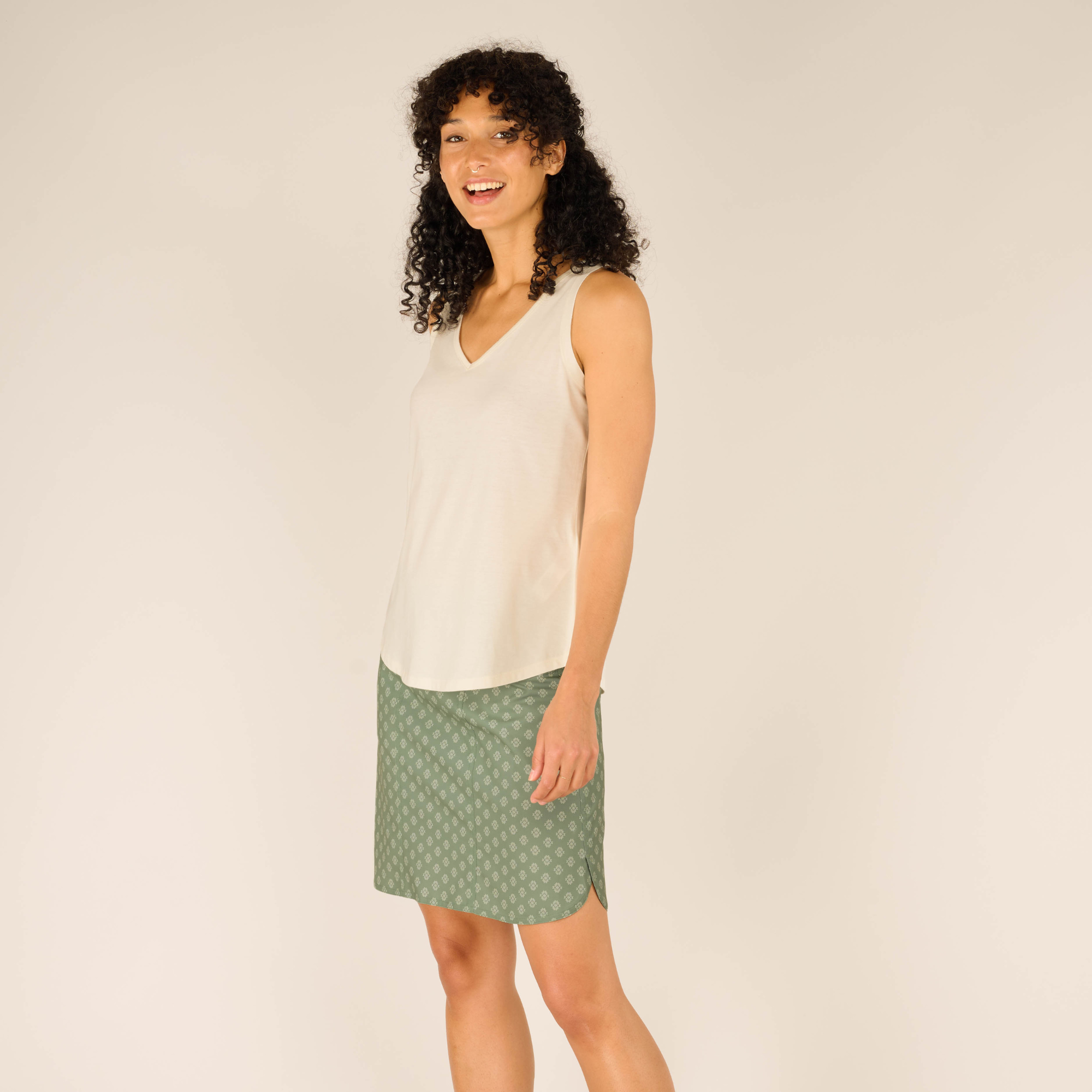 A female model with curly dark hair stands in profile, wearing a Sherpa Adventure Gear Varuna V-Neck Tank in Cream and a green knee-length skirt with a subtle repeating pattern. She is smiling, looking toward the camera with her left arm resting at her side. The tank top drapes loosely, and the fabric appears soft. The background is a plain, neutral beige.