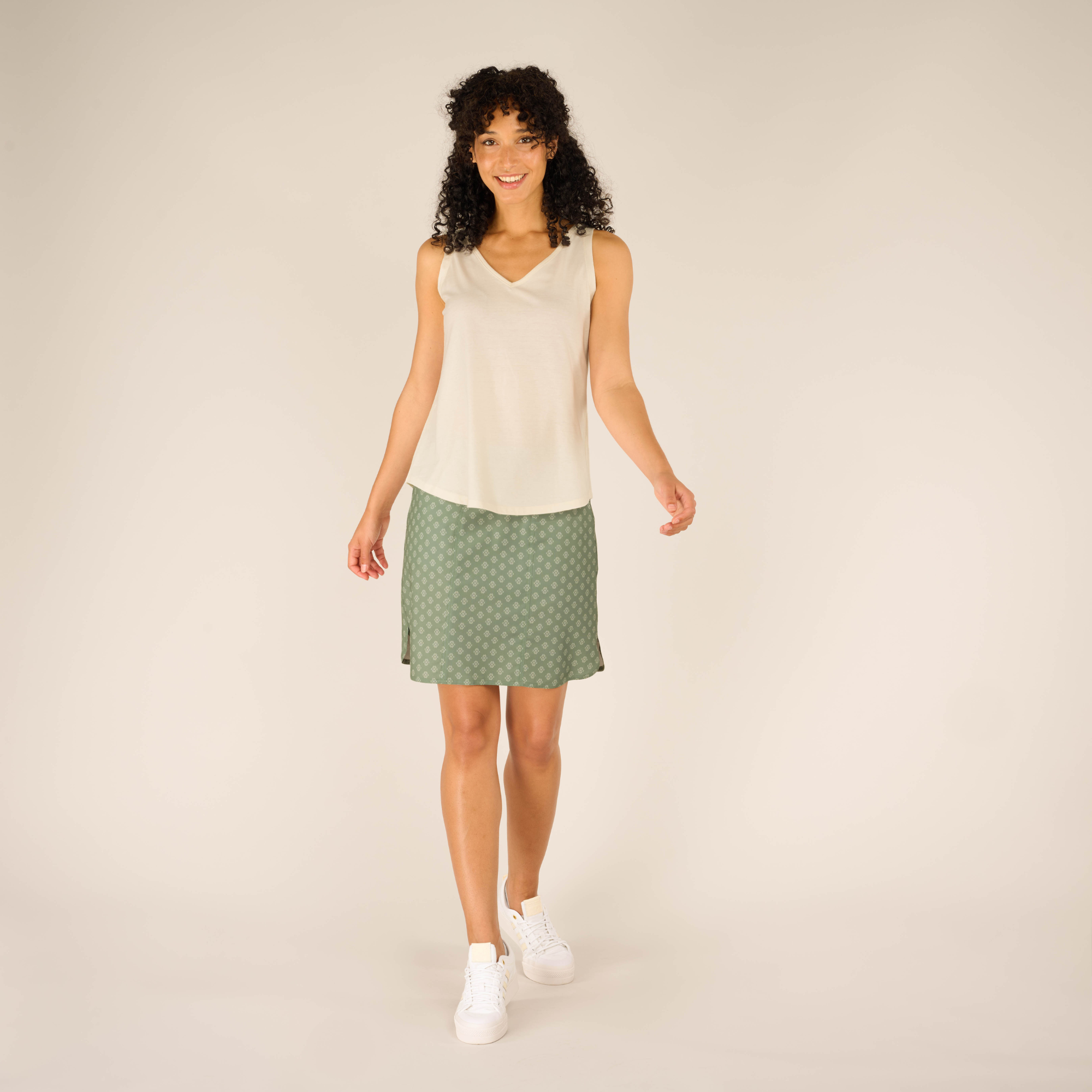 The model stands facing the camera, smiling brightly while wearing the Sherpa Adventure Gear Varuna V-Neck Tank in Cream and green patterned skirt. She is standing with her arms slightly out to her sides, and her feet slightly apart, showing the full length of the skirt. She wears white sneakers. The setting remains neutral and minimalistic.