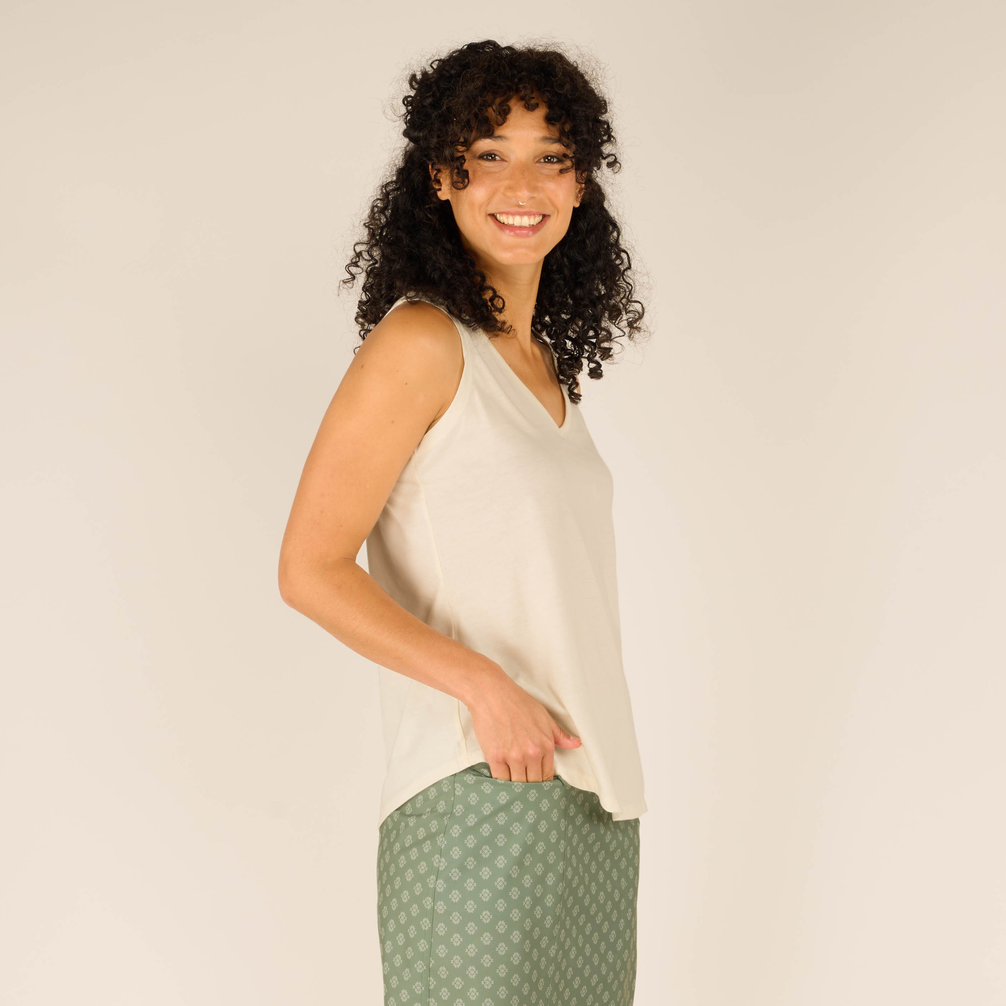 Close-up side view of the model in the Sherpa Adventure Gear Varuna V-Neck Tank in Cream, showing the smooth fabric and loose fit. She places her hand in the pocket of the green skirt, highlighting the functional detail of the skirt. The V-neckline is visible, and the top’s hemline slightly curves.