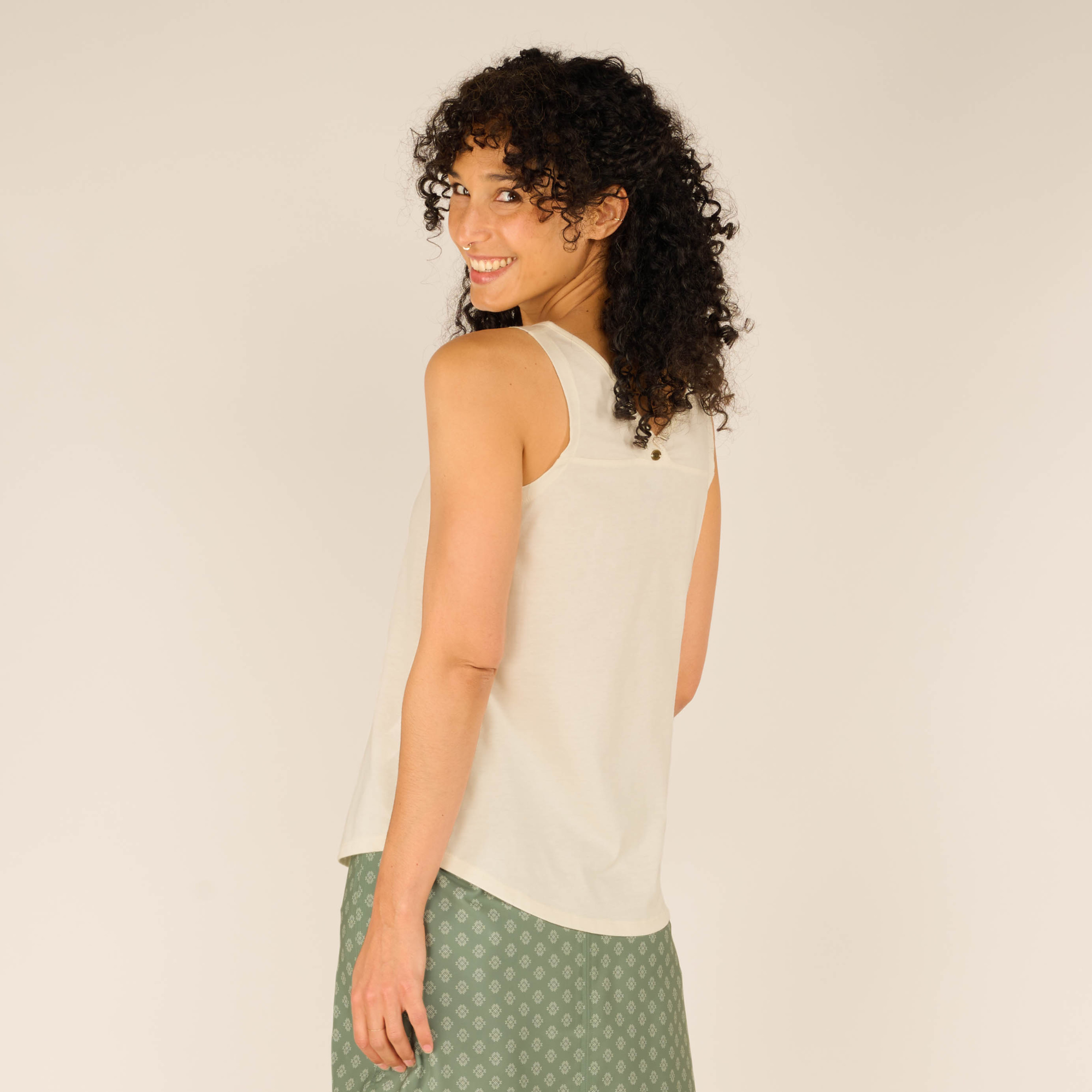 Back view of the model wearing the Sherpa Adventure Gear Varuna V-Neck Tank in Cream and green skirt. She turns her head slightly toward the camera, smiling. The back of the tank top has a small decorative button detail just below the neckline. The armholes appear comfortably cut, and the back hemline mirrors the front's gentle curve.