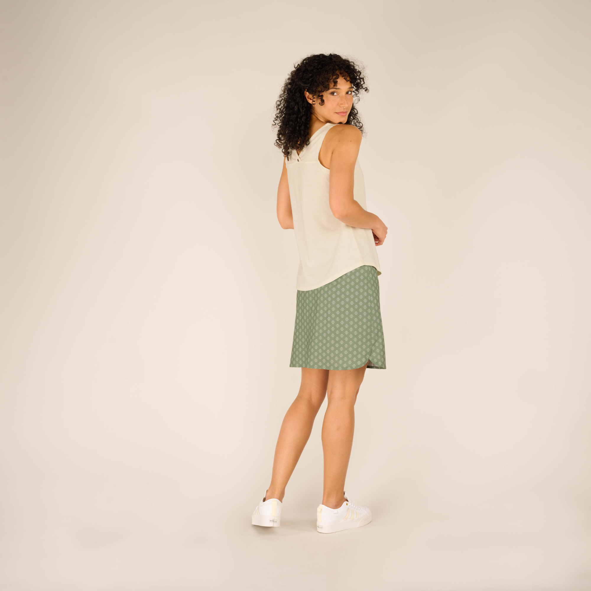 The model stands turned slightly away from the camera, showing the back and side view of the outfit. The Sherpa Adventure Gear Varuna V-Neck Tank in Cream flows smoothly over the waistband of the green skirt, which features a slight slit on the side. The model’s white sneakers add a casual touch.