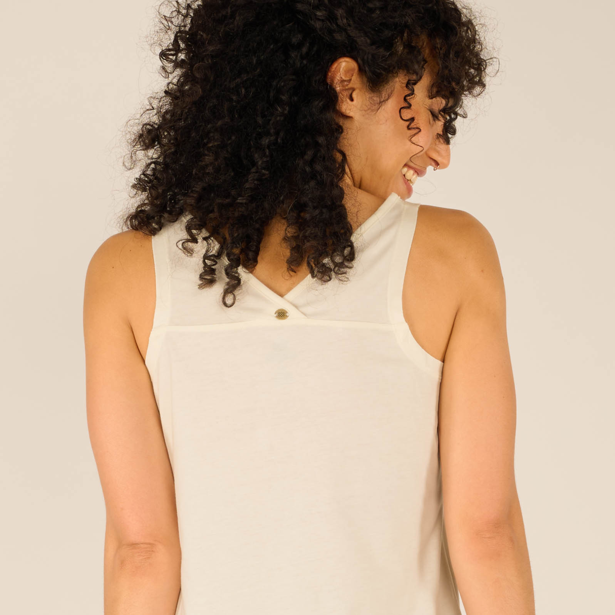 Close-up of the upper back of the Sherpa Adventure Gear Varuna V-Neck Tank in Cream, focusing on the small button embellishment centered below the neckline. The fabric’s soft texture is visible, and the model’s curly hair frames the top. The shoulder seams and armhole finish appear clean and well-constructed.