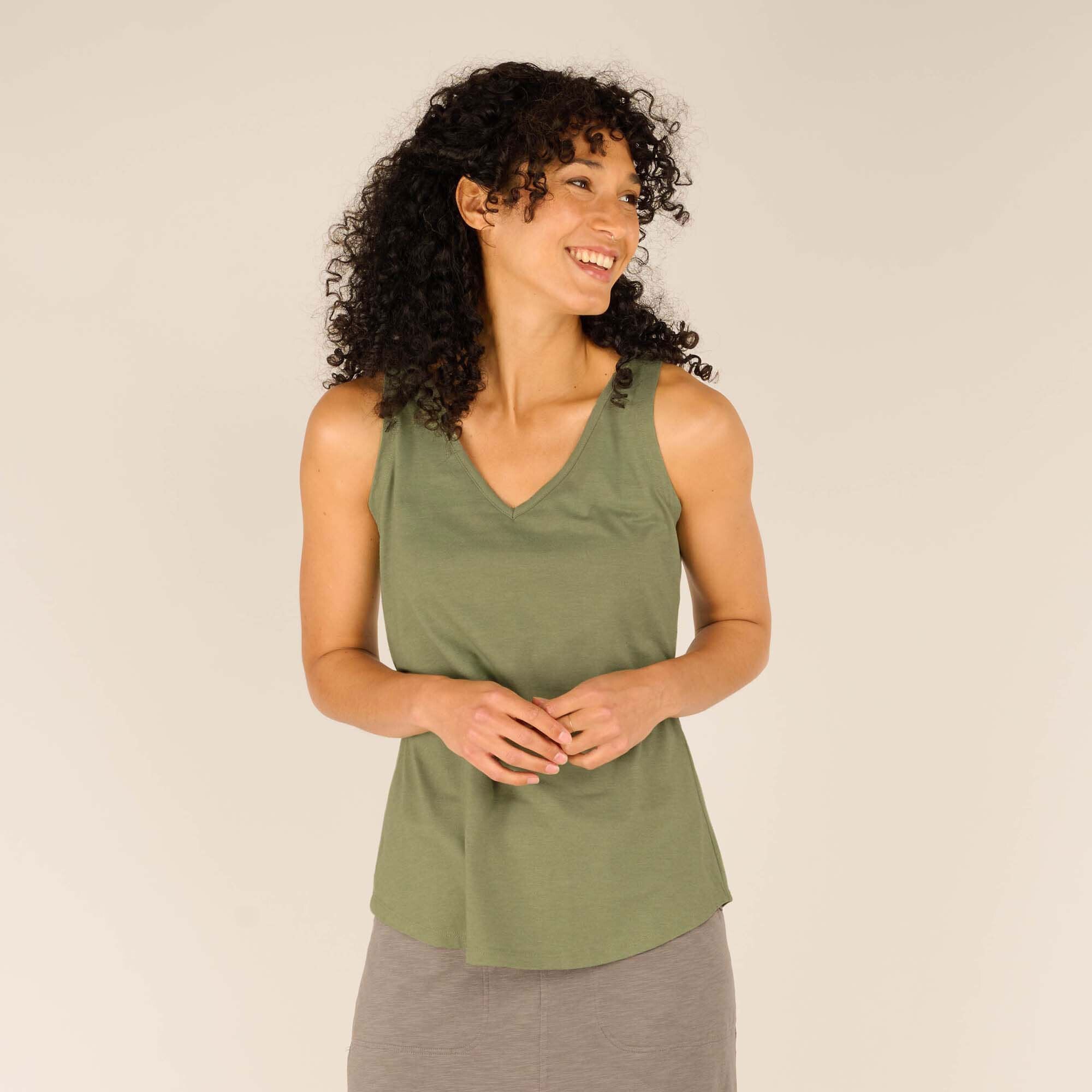 A female model with curly dark hair smiles while wearing a Sherpa Adventure Gear Varuna V-Neck Tank in Green. The sleeveless top features a flattering V-neckline and a slightly curved hemline. The model pairs the tank with a grey casual skirt and stands against a neutral beige backdrop, hands gently clasped together in front of her.