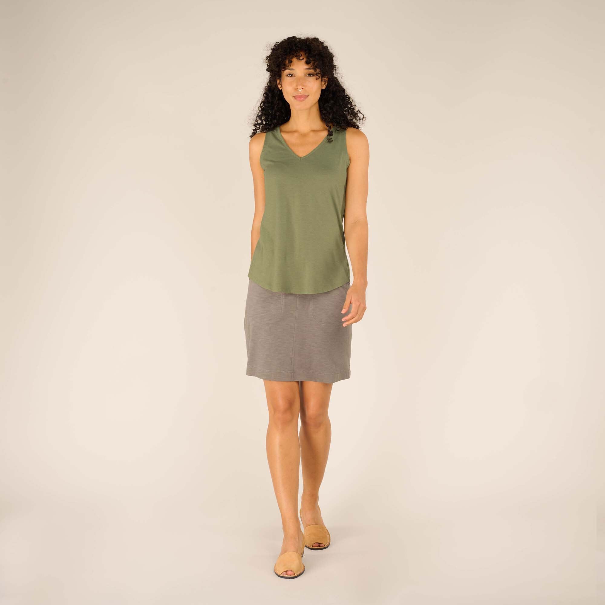 Full-body view of the female model wearing the Sherpa Adventure Gear Varuna V-Neck Tank in Green. The model walks toward the camera, showcasing the tank's relaxed fit and length, which falls just below the waist. The grey skirt and tan slip-on shoes complete her comfortable, casual look.