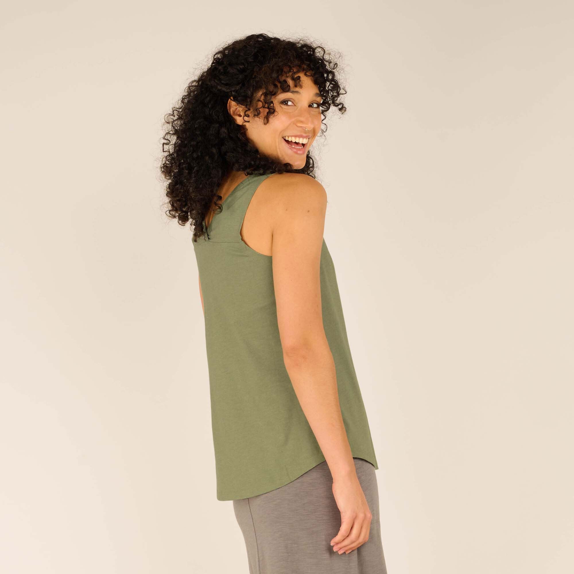 Side view of the female model wearing the Sherpa Adventure Gear Varuna V-Neck Tank in Green. She looks over her shoulder with a playful smile, highlighting the tank's clean side seams and sleeveless cut, perfect for warm weather. The fabric drapes softly around her torso.