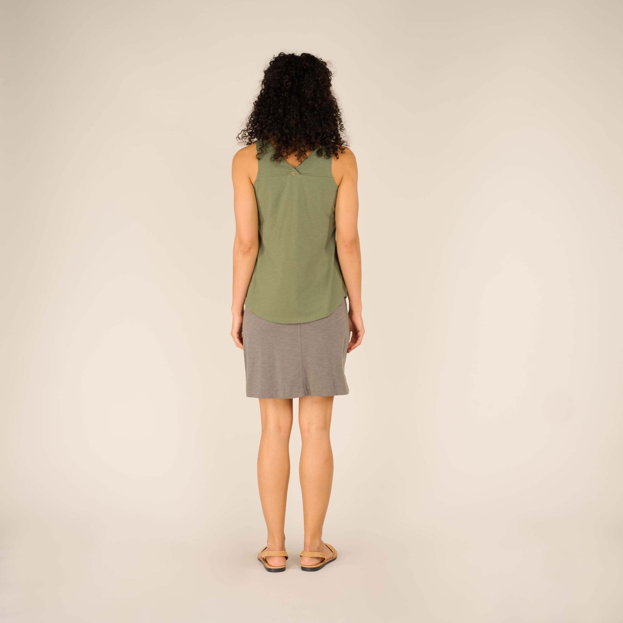 Back view of the female model wearing the Sherpa Adventure Gear Varuna V-Neck Tank in Green. The back neckline has a subtle V-shape detail with a small decorative button at the center, adding a touch of interest to the otherwise simple design. The tank flows over her grey skirt.