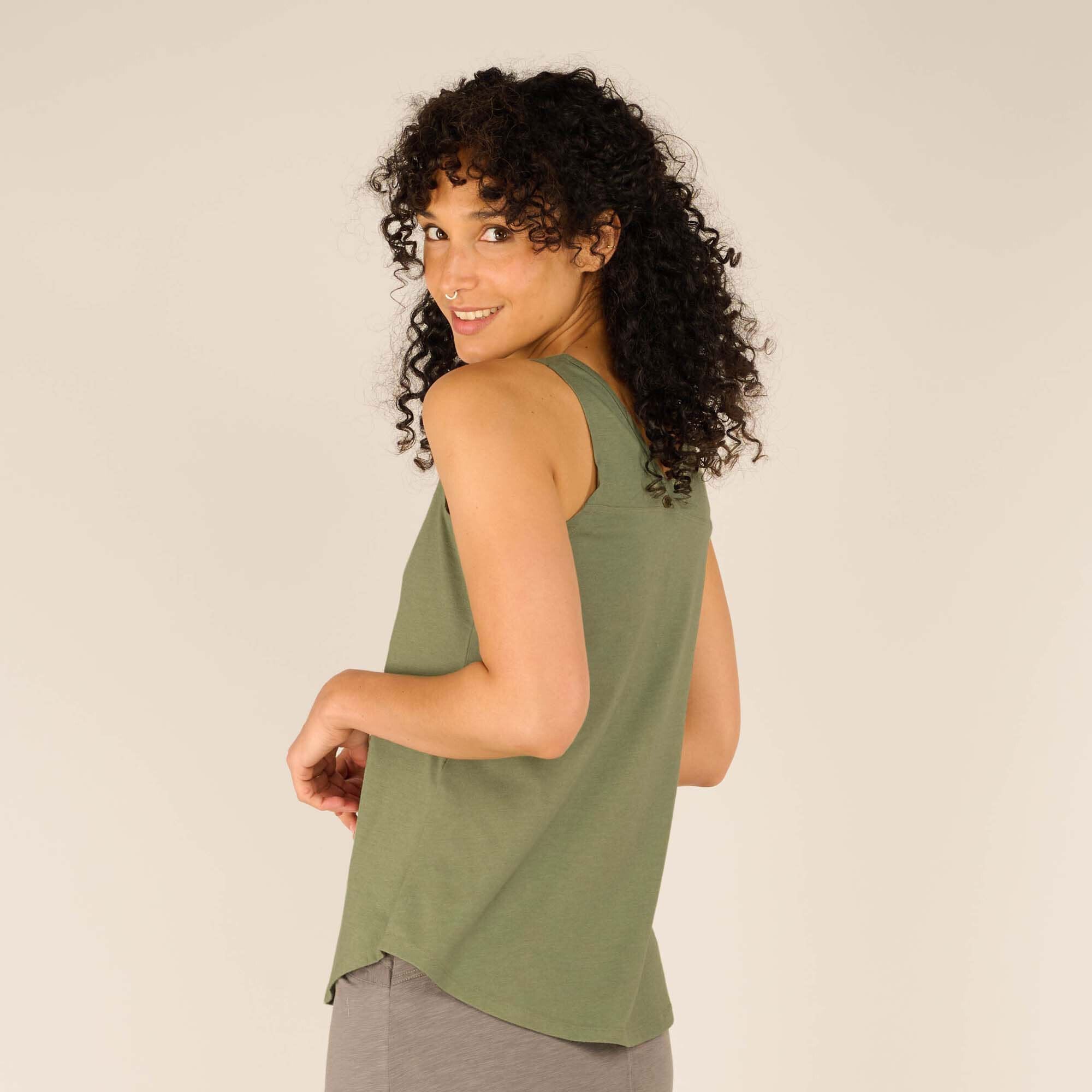 Close-up side view of the female model wearing the Sherpa Adventure Gear Varuna V-Neck Tank in Green. She gently turns toward the camera with a soft smile, showing the smooth fabric and clean lines of the top. The green shade complements her skin tone and pairs well with the grey skirt.