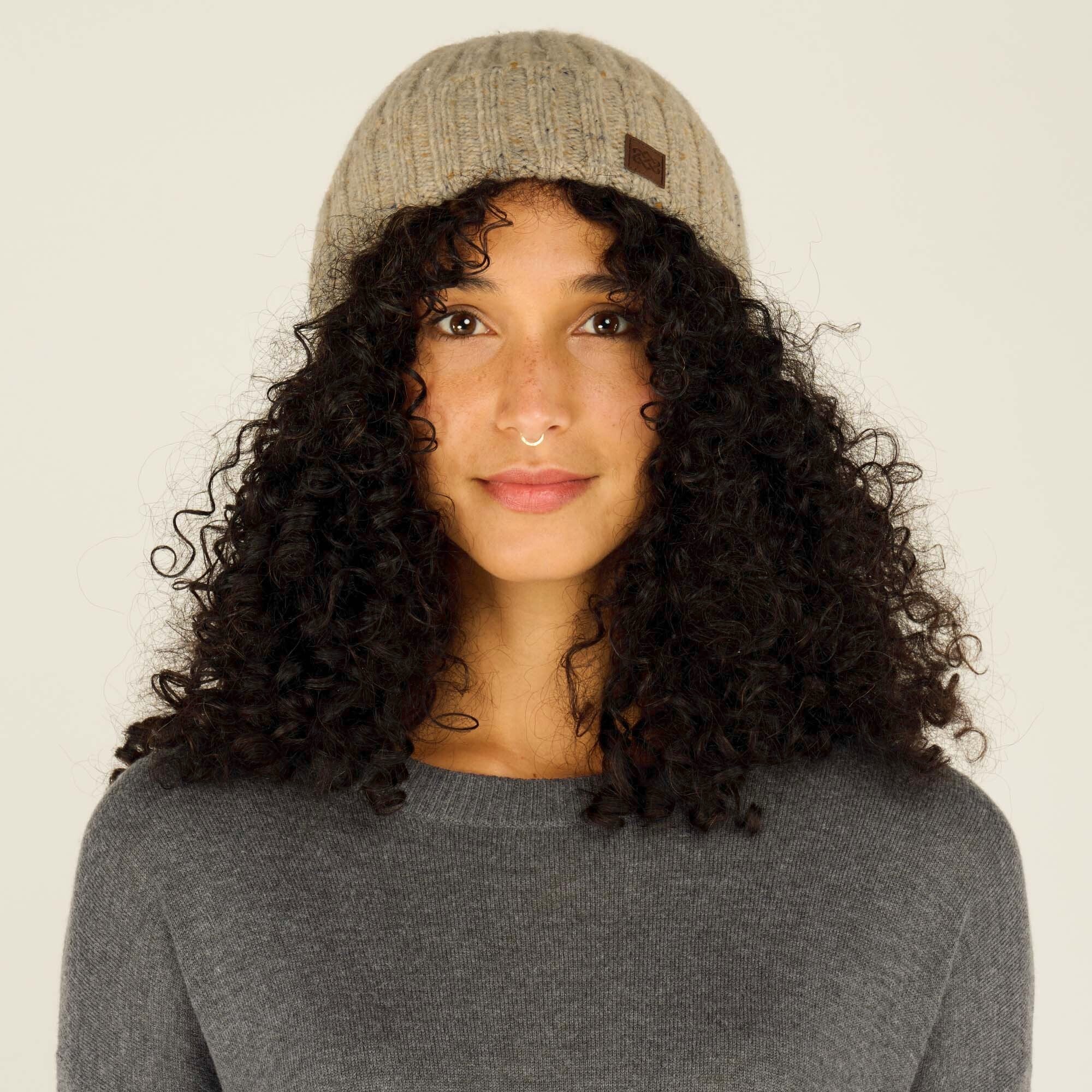 A model with curly black hair is wearing a Sherpa Adventure Gear Varuni Classic Beanie in Beige with subtle speckles. The beanie features a small rectangular leather tag with a logo on the left side. The model wears a gray sweater and gazes directly at the camera, with a neutral background highlighting the details of the hat.