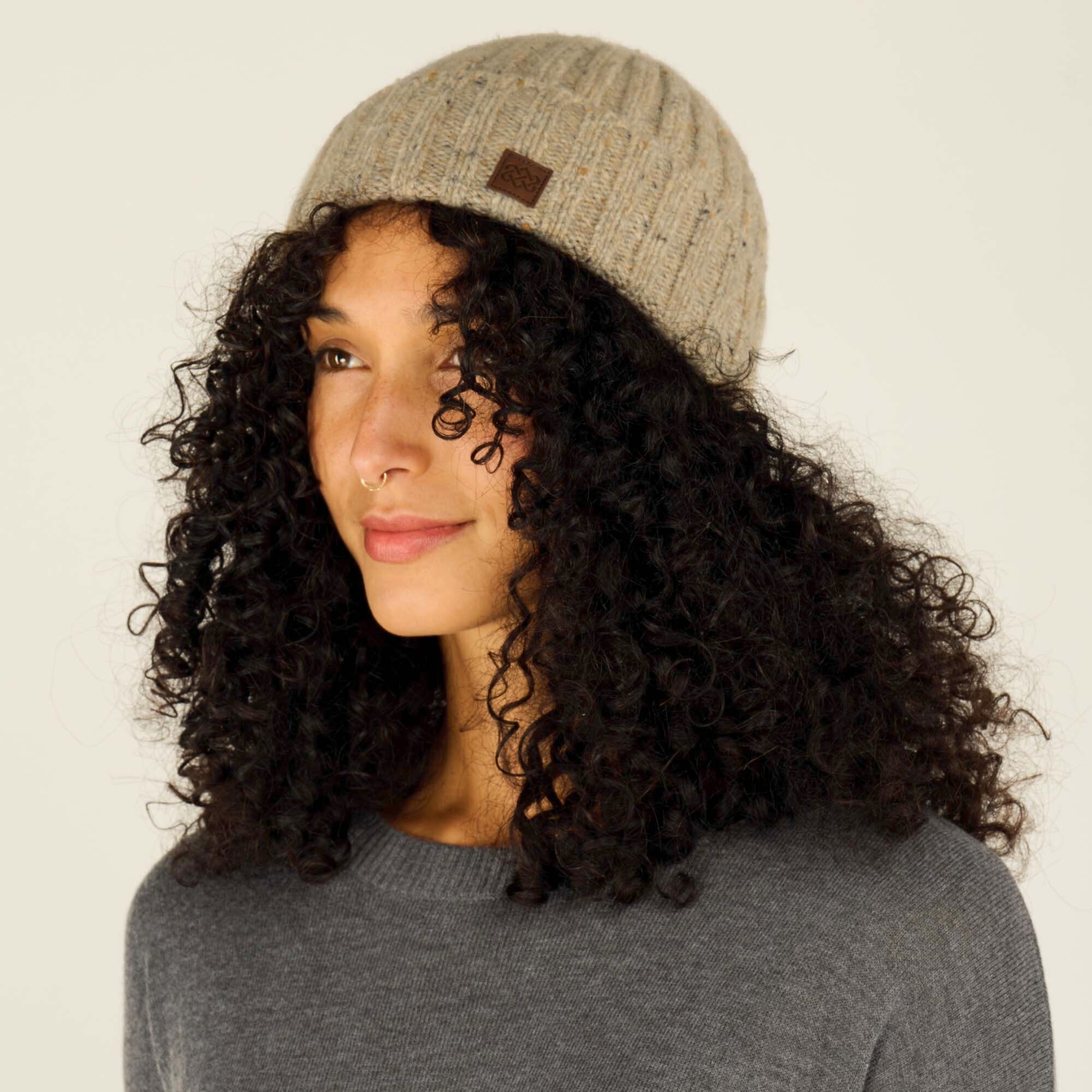 A side view of the same Sherpa Adventure Gear Varuni Classic Beanie in Beige on the model, showcasing the rounded fit and subtle speckled texture. The small leather logo tag is visible on the side of the beanie. The model is smiling softly and looking off into the distance, with her curly black hair peeking out slightly under the beanie. The neutral background emphasises the beanie's classic design.