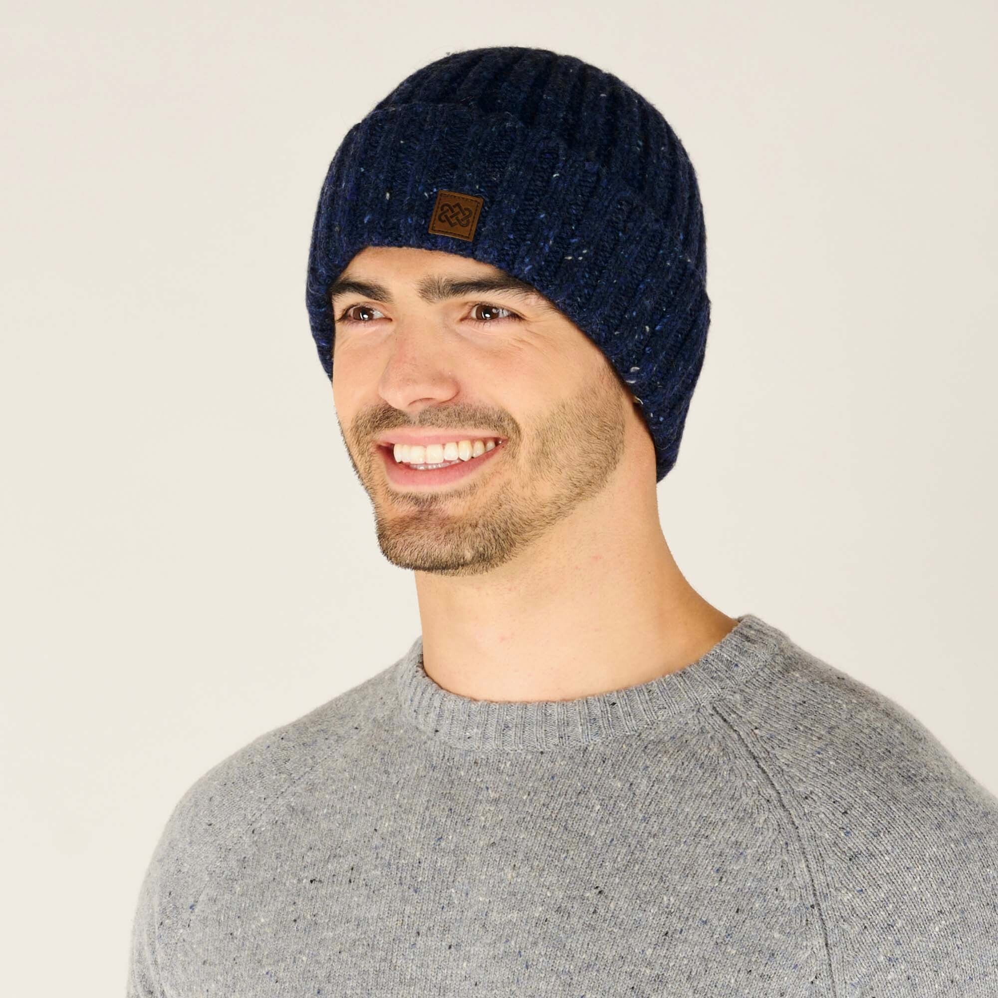 A side view of the same Sherpa Adventure Gear Varuni Classic Beanie in Blue, showcasing its snug fit and textured knit pattern. The brown leather patch with an embossed knot design is visible on the side. The model is slightly turned, smiling, and the light grey sweater provides a neutral backdrop, highlighting the beanie’s classic and versatile design.