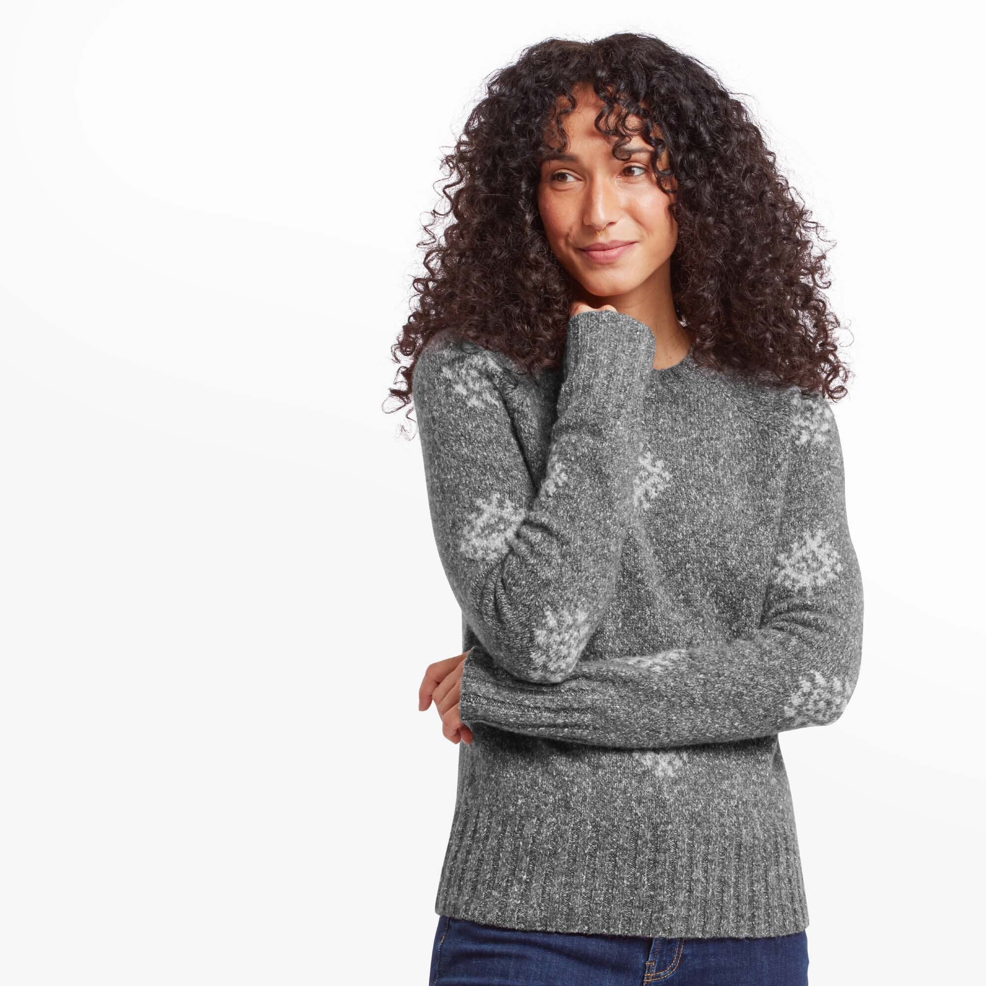 A woman with curly hair is wearing a Sherpa Adventure Gear Vindo Crew in Grey with light grey geometric snowflake patterns. She is slightly leaning her head on her hand, smiling gently. The soft knit texture of the sweater and the casual yet cozy design are prominently featured.