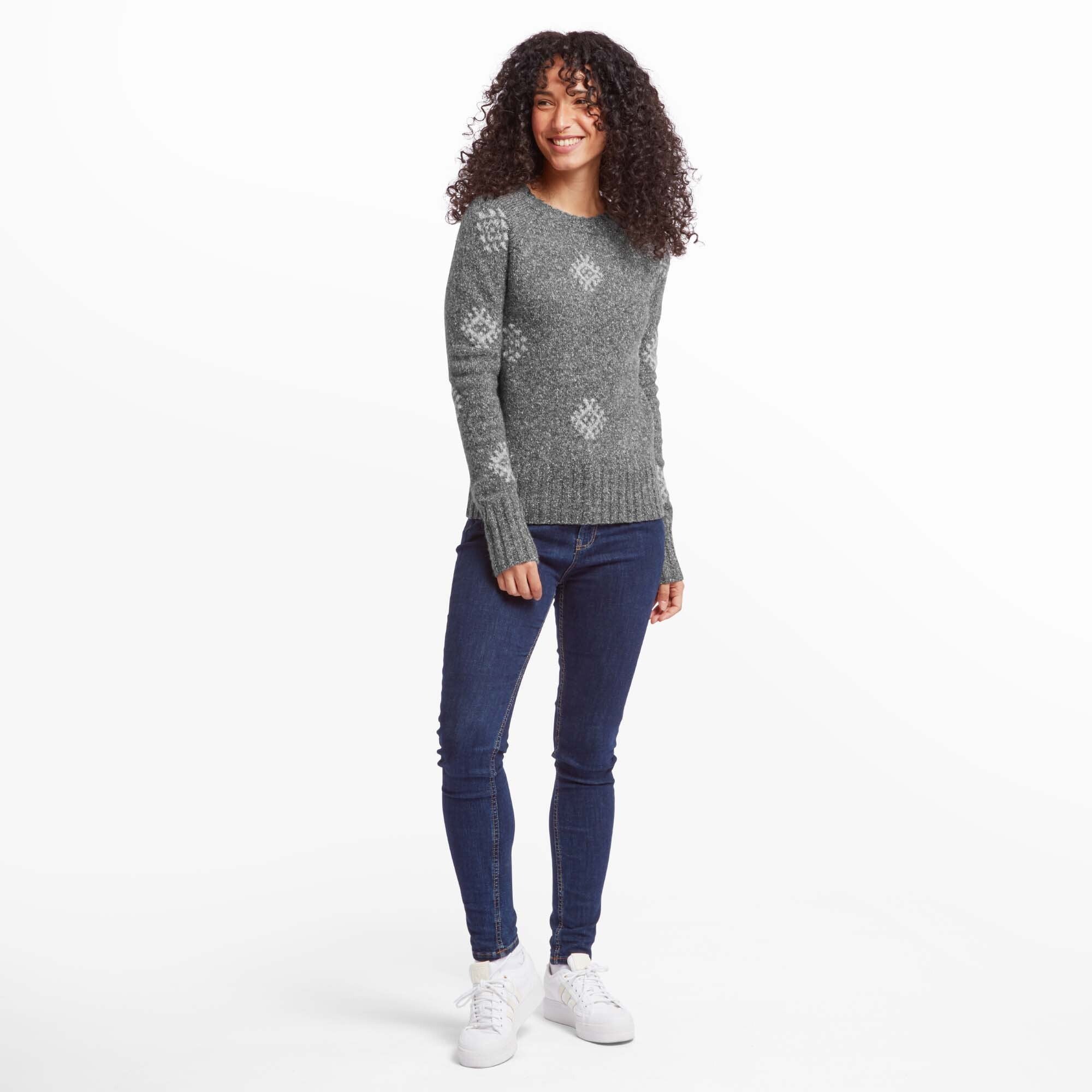 A full-body view of the same woman, styled in the Sherpa Adventure Gear Vindo Crew in Grey paired with dark blue skinny jeans and white trainers. She is standing with a relaxed posture and a cheerful expression, highlighting the versatile and comfortable look of the outfit.