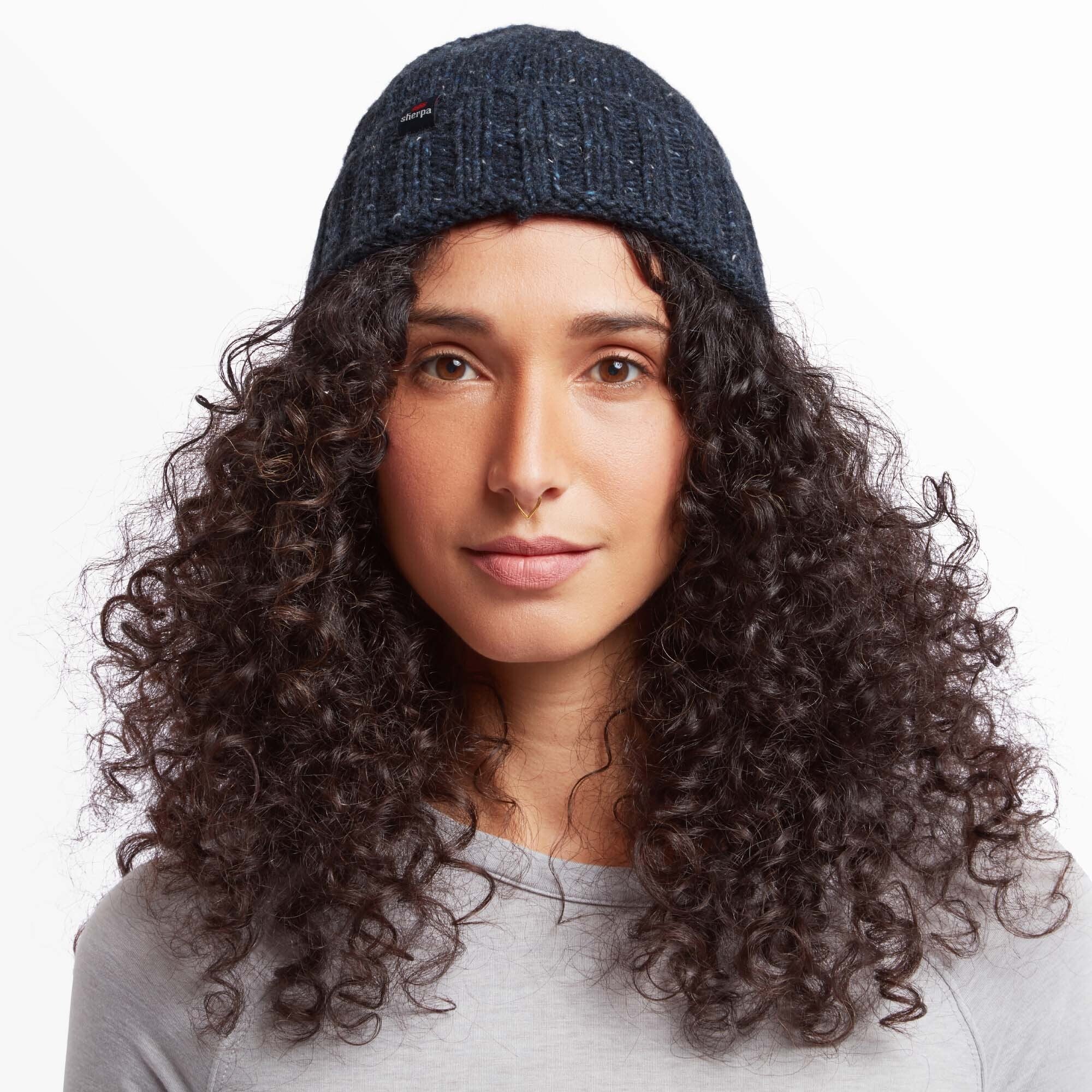 A close-up front view of a person wearing a Sherpa Adventure Gear Vishnu Hat in Blue with subtle fleck detailing. The hat has a small label with the Sherpa Adventure Gear logo on the front. The person has voluminous curly hair cascading around the hat and is wearing a light gray top. Their facial expression is calm and composed.