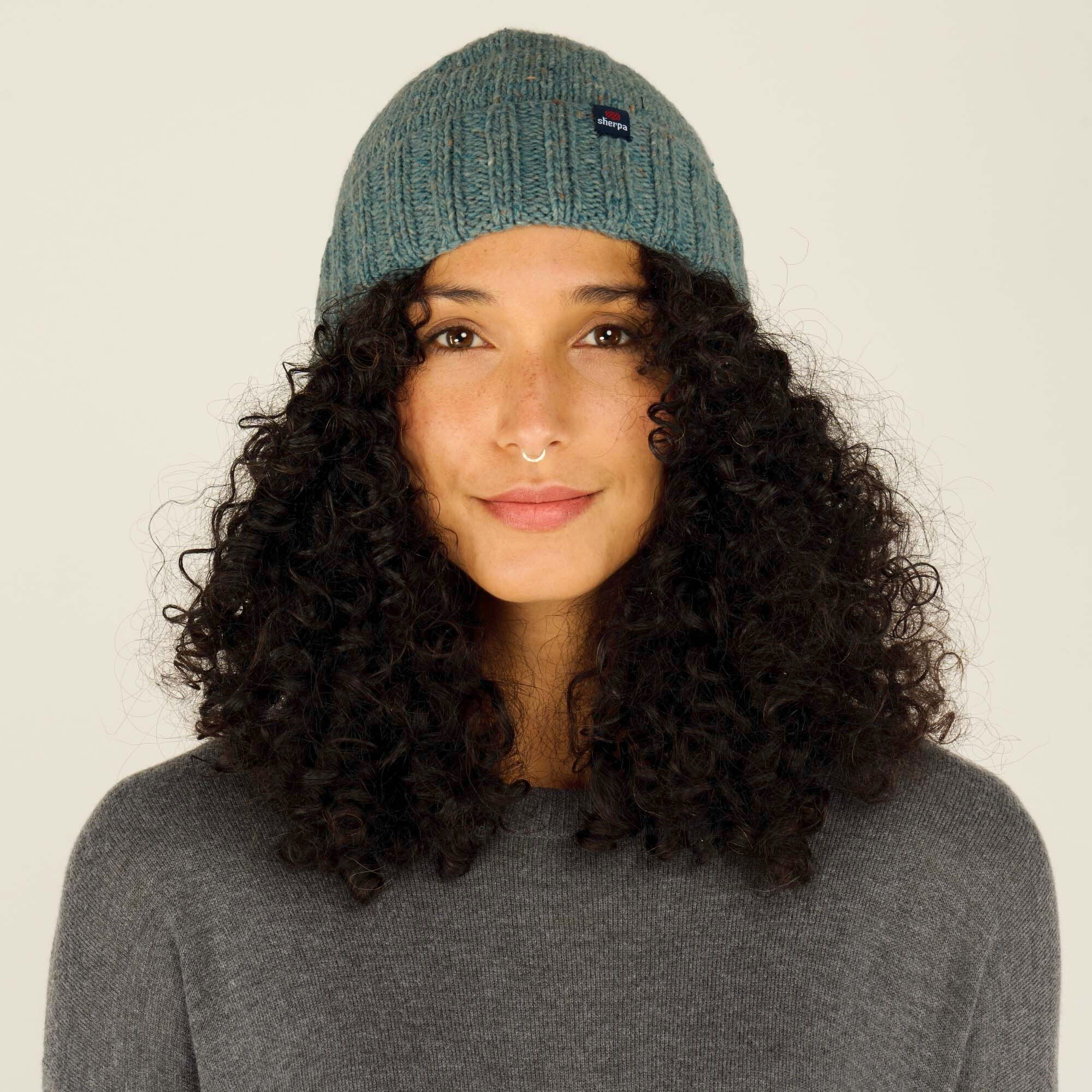 The image showcases a woman wearing a Sherpa Adventure Gear Vishnu Hat in Blue. Her curly black hair frames her face, and the hat features a subtle flecked pattern with a small logo patch near the rim. She is wearing a gray sweater, looking directly into the camera with a soft smile, and the background is neutral.