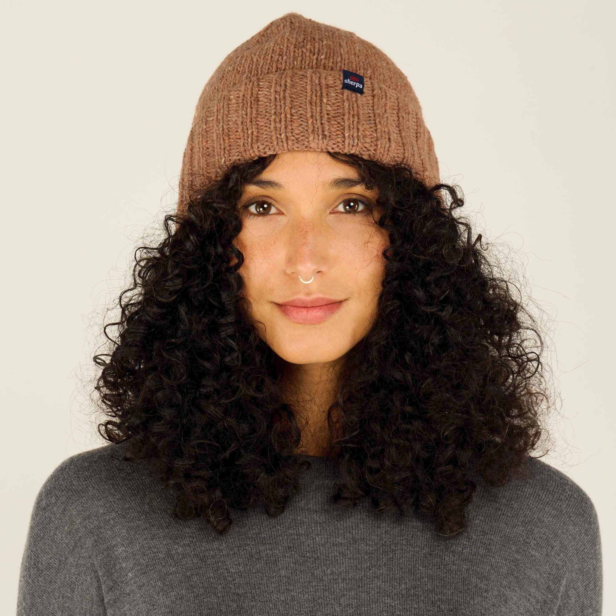 A close-up front view of a woman wearing a cozy Sherpa Adventure Gear Vishnu Hat in Orange. The hat has a ribbed design with a small Sherpa logo patch on the front. She has curly black hair cascading around her face, is dressed in a grey sweater, and is looking directly at the camera with a subtle smile.