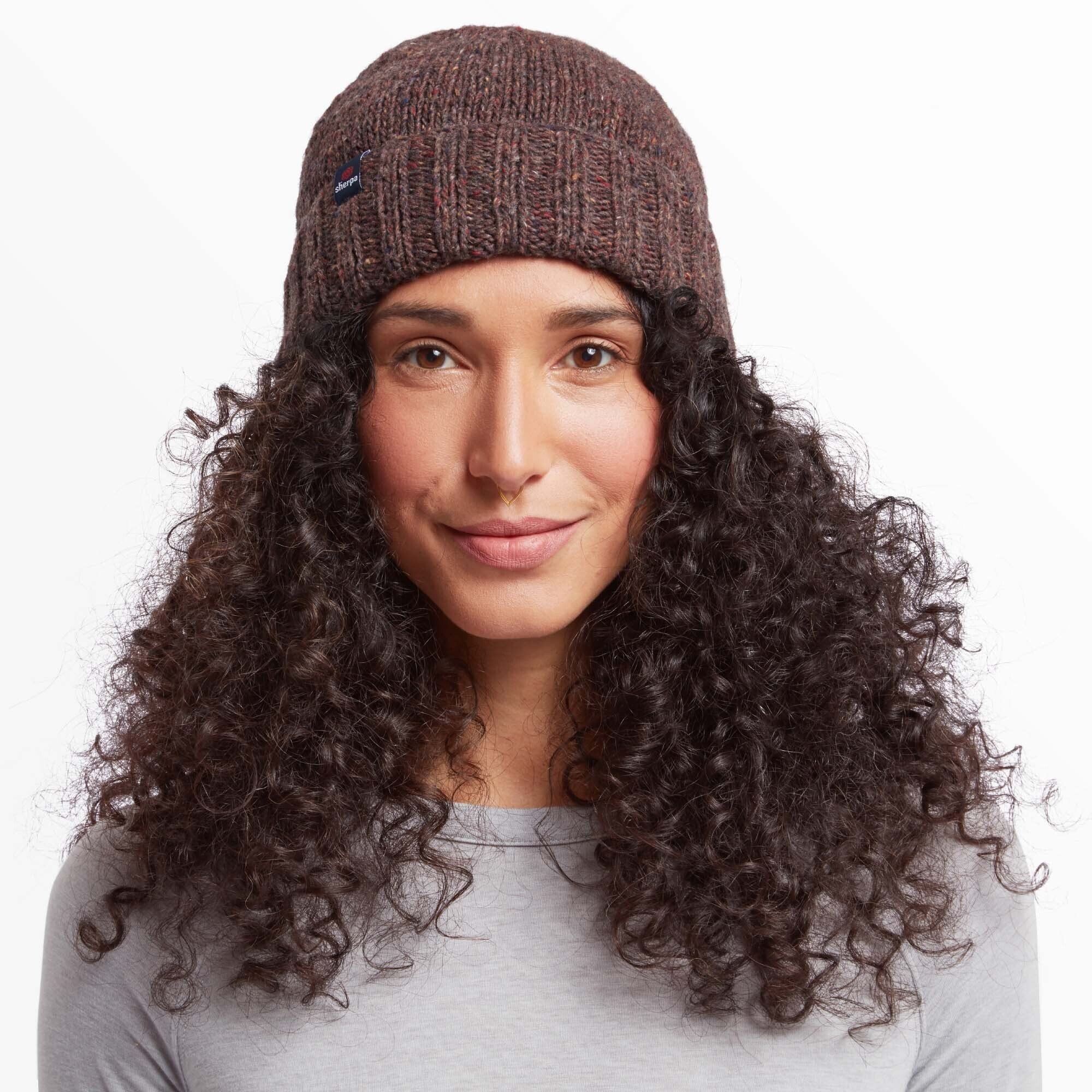 Close-up front view of a woman wearing a Sherpa Adventure Gear Vishnu Hat in Red with a ribbed fold-over brim. The hat features a small Sherpa Adventure Gear logo tag on the side. The model has voluminous curly hair, a soft smile, and is dressed in a light gray top. The background is plain white, emphasising the hat's details.