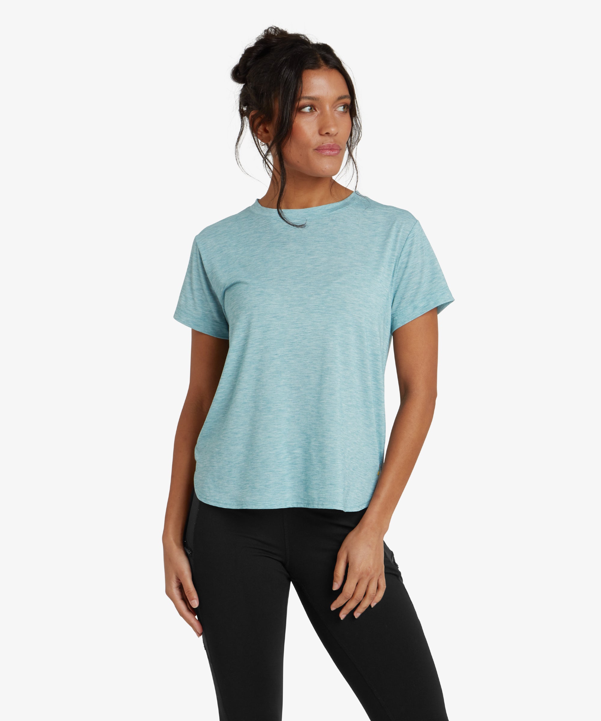 A woman is wearing a Sherpa Adventure Gear Asha Crew in Blue with short sleeves. The T-shirt has a relaxed fit and a slightly curved hemline. The material appears soft and lightweight, making it ideal for casual or active wear. The model is smiling and has one hand resting on her hip, showcasing the comfortable fit.