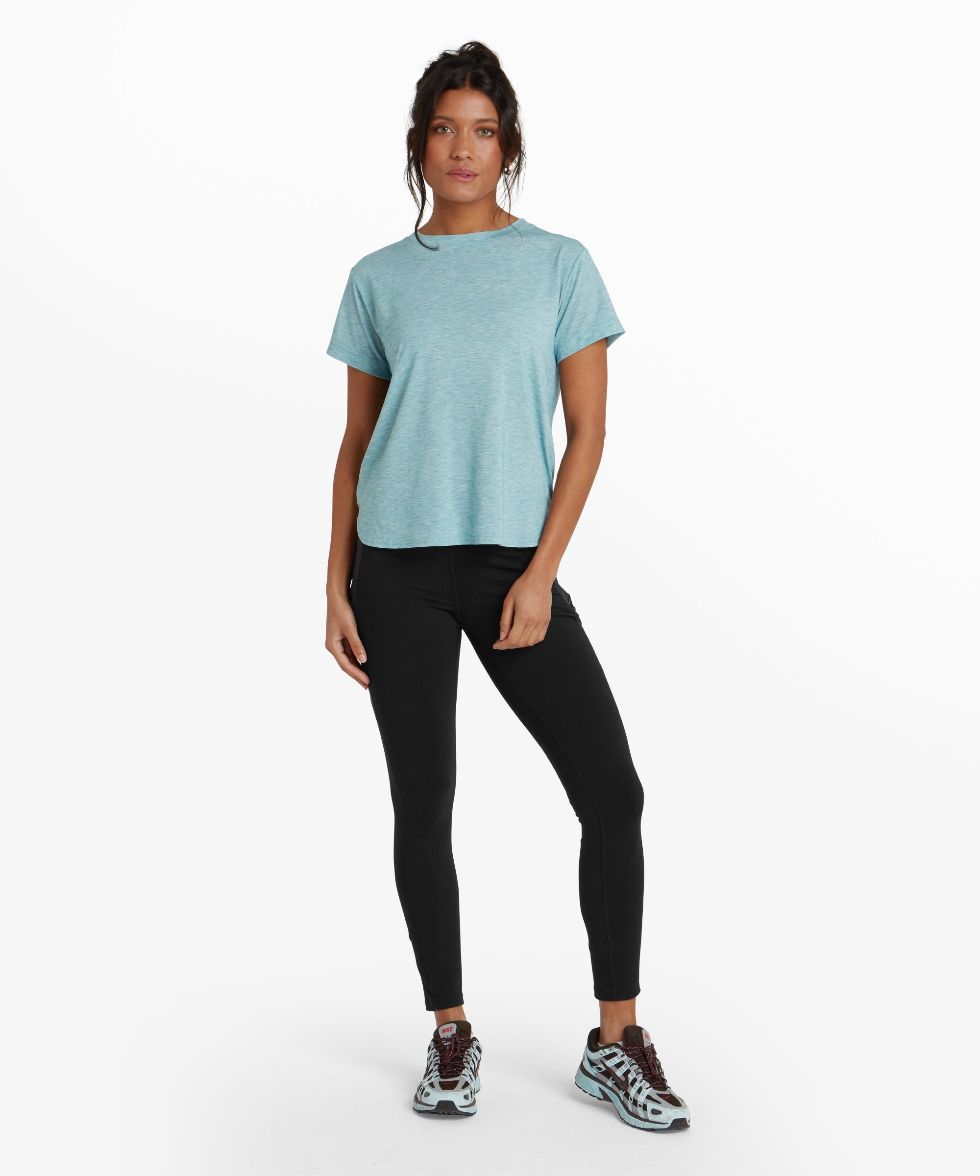 The same woman is shown from a side angle, highlighting the Sherpa Adventure Gear Asha Crew in Blue's flattering fit along her torso and the extended back hem. The curved hemline creates a casual, laid-back look, while the short sleeves and crew neckline maintain a classic style. Her slightly turned pose emphasises the shirt's versatile cut.